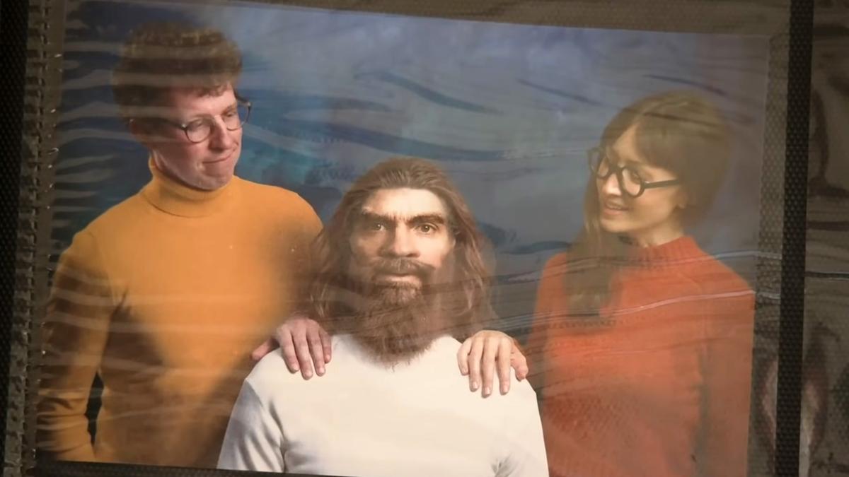 A man and a woman smiling with their hands on the shoulders of a Neanderthal figure with long hair and a beard but in modern clothes.