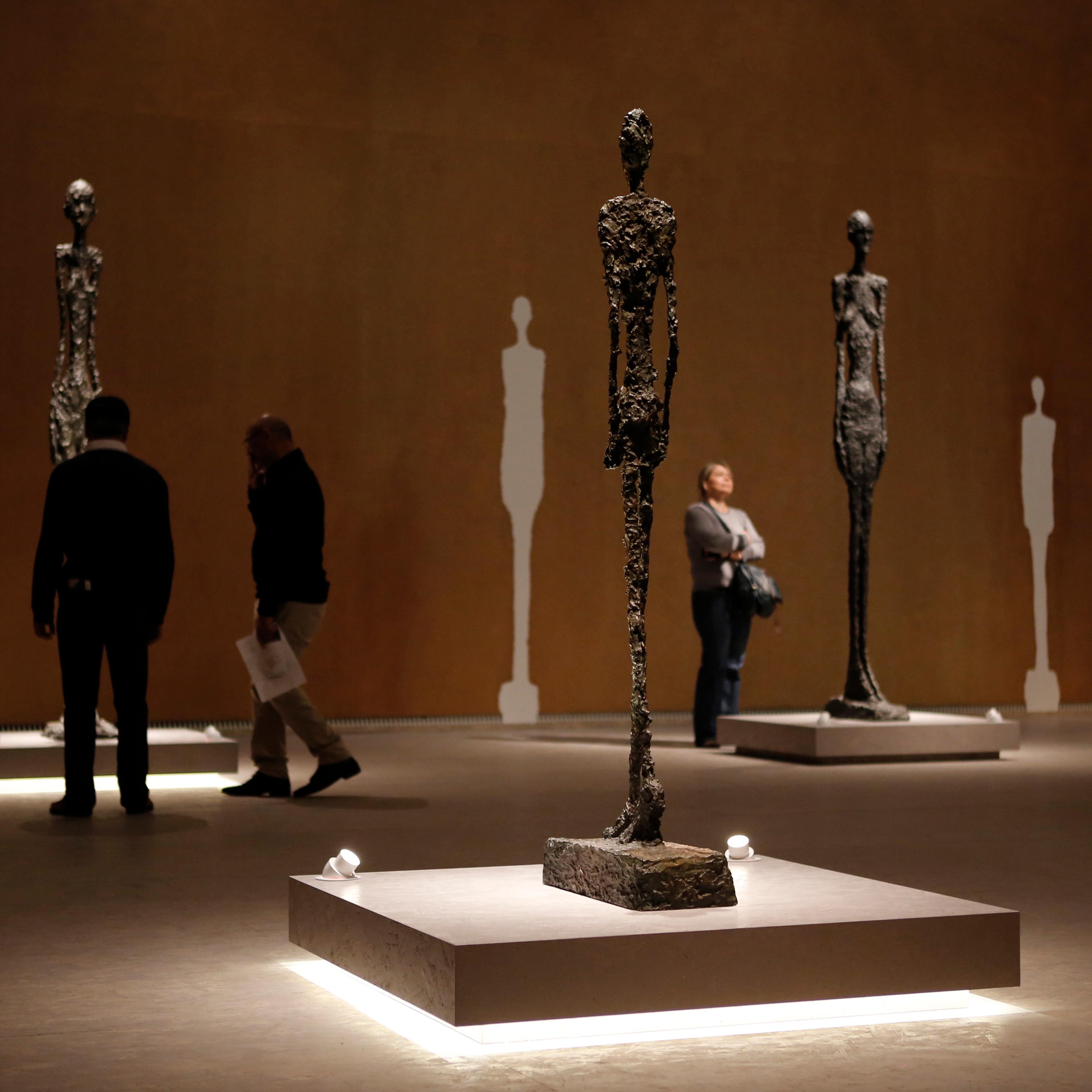 Photo of people viewing tall abstract sculptures in a dimly lit gallery with silhouetted artwork on the walls.