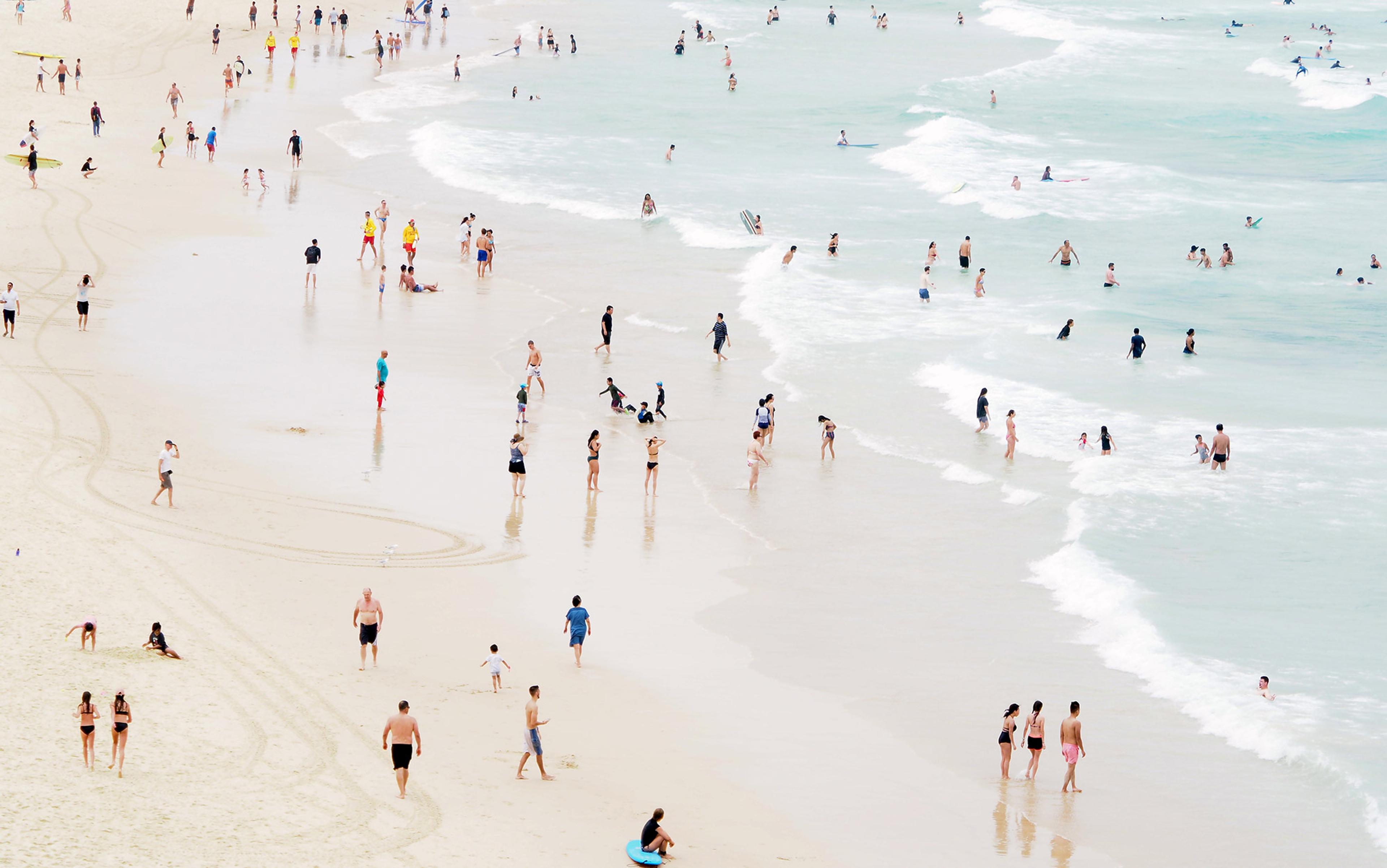 A crowded beach with people swimming, walking on the shore and playing in the sand. The sea has gentle waves.