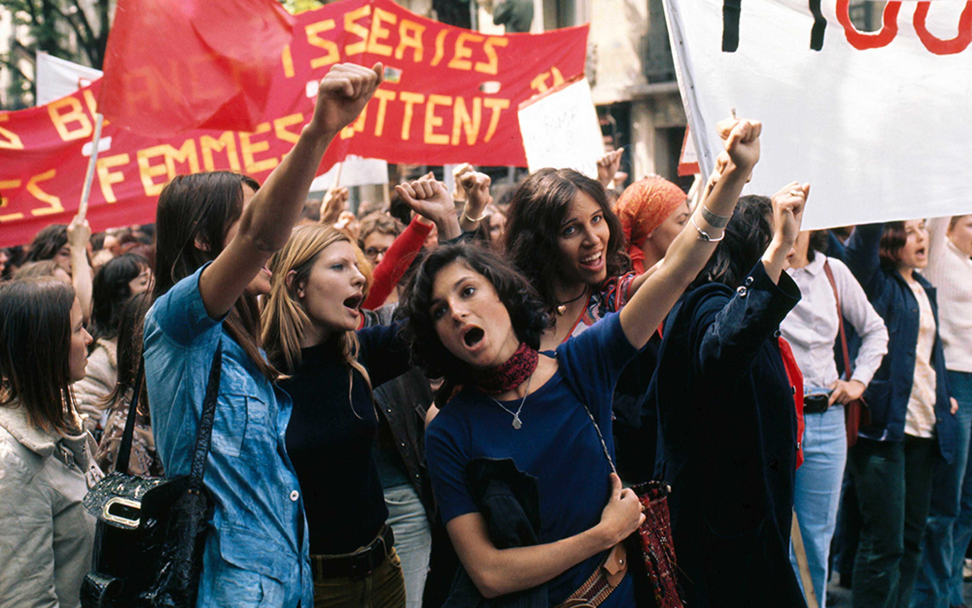 A group of women protesting, holding red and white banners, fists raised, some shouting, in an outdoor urban setting.