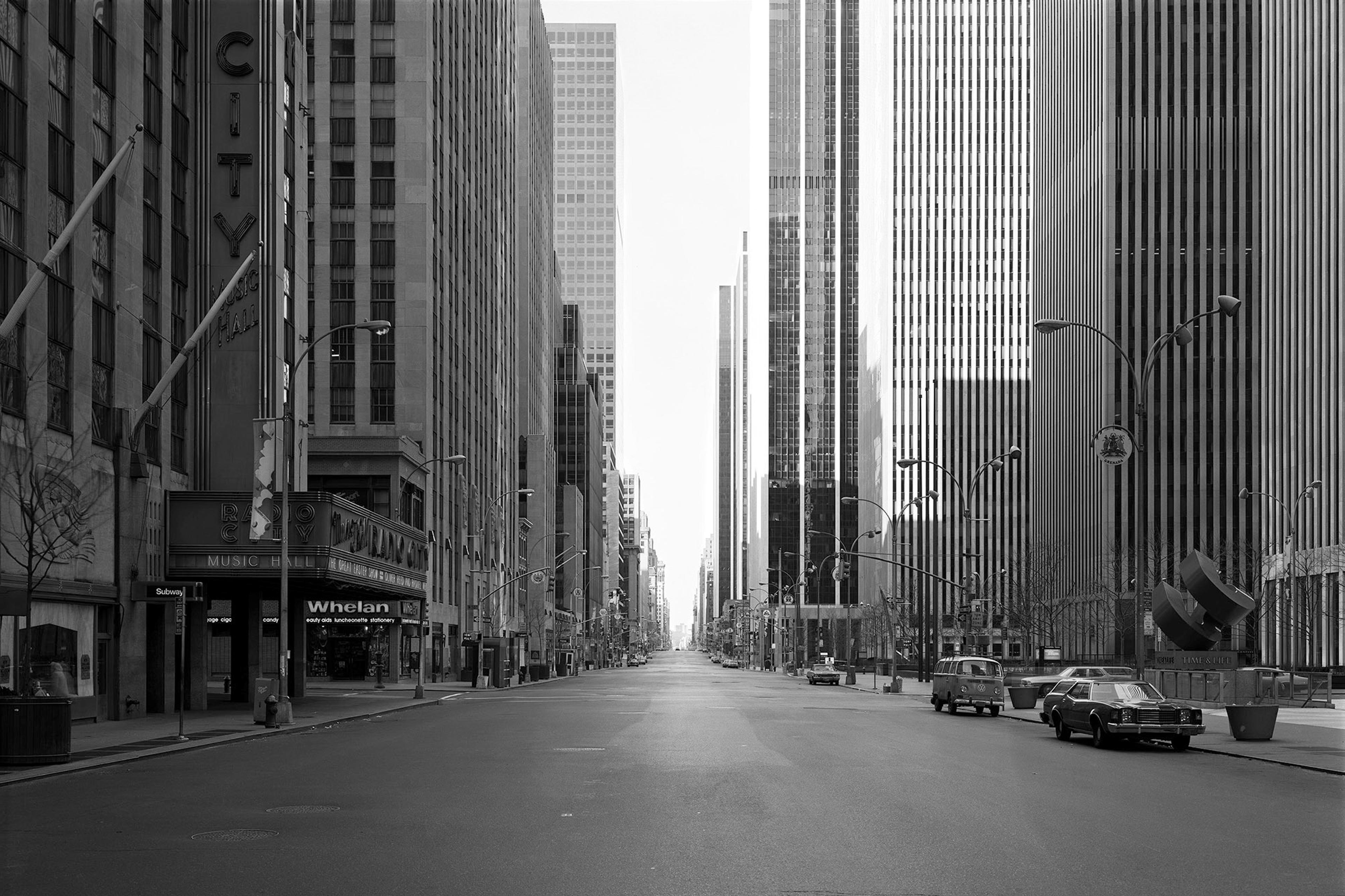Black and white photo of an empty city street flanked by tall buildings, featuring vintage cars and a distant vanishing point.