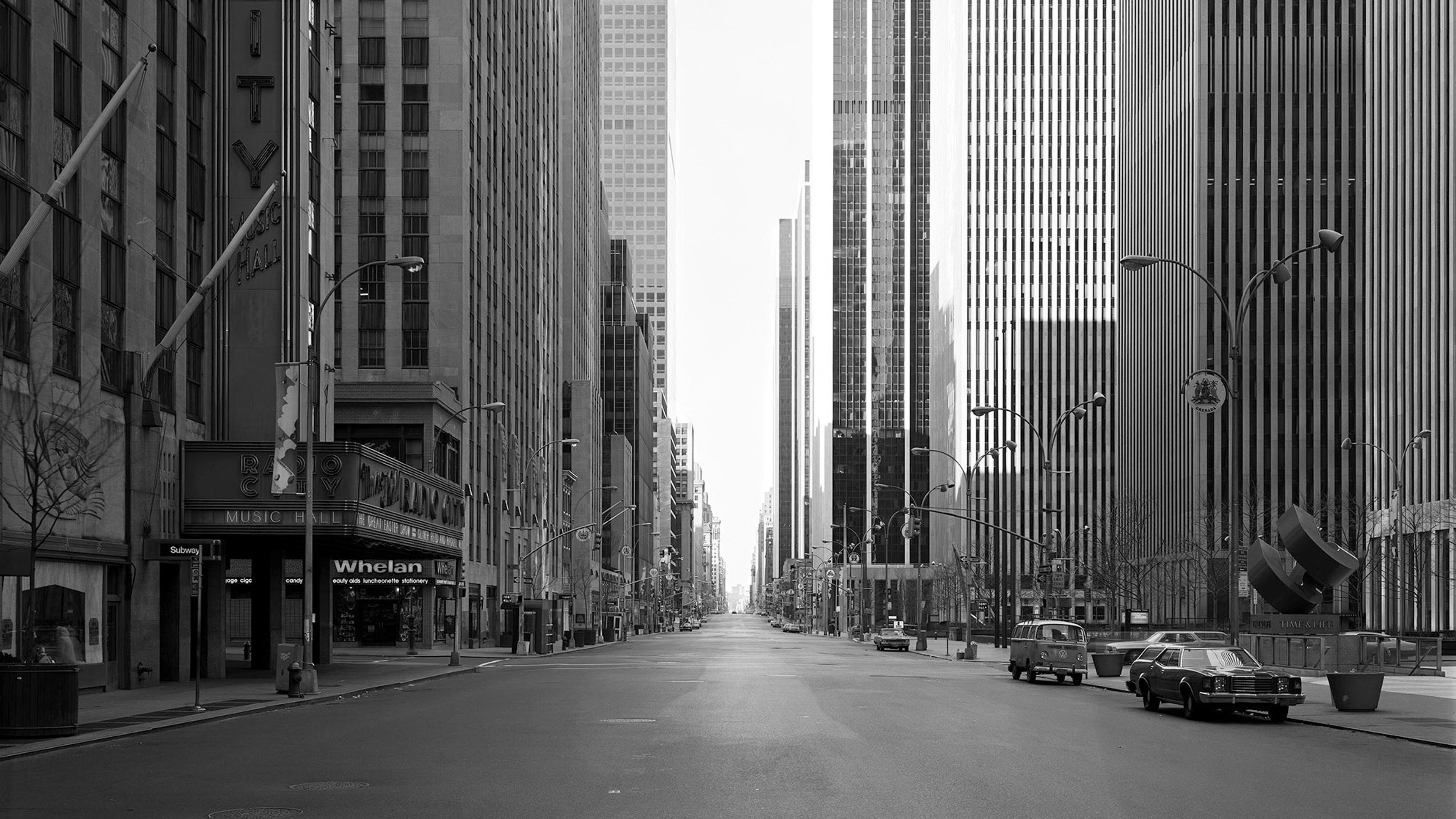 Black and white photo of an empty city street flanked by tall buildings, featuring vintage cars and a distant vanishing point.