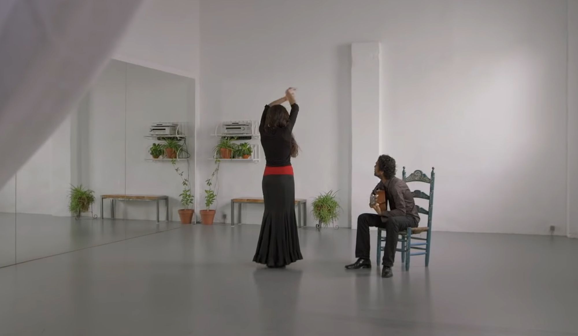 A dancer in a black dress and red sash dances in front of a guitarist seated on a chair, in a studio with mirrors and plants.