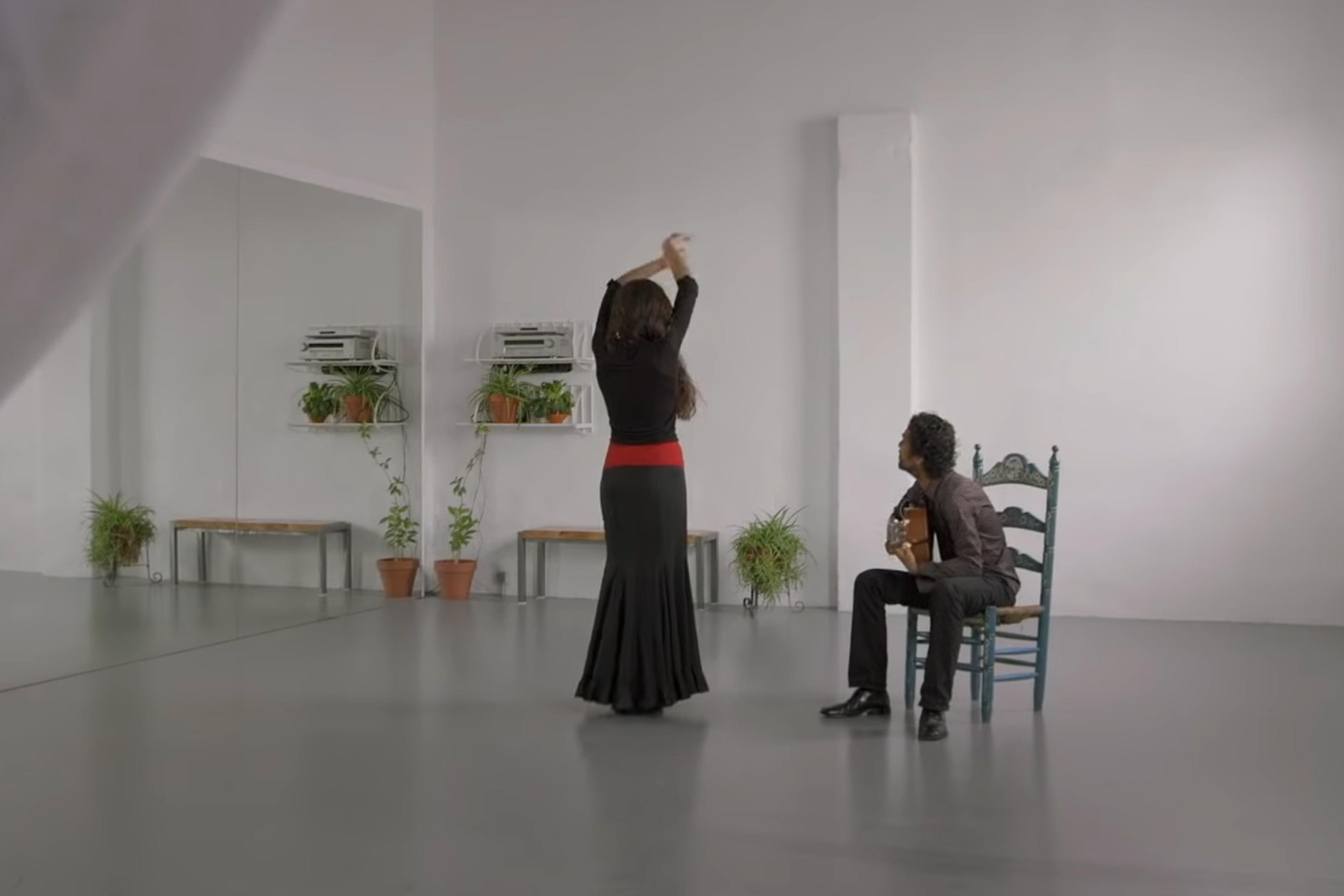 A dancer in a black dress and red sash dances in front of a guitarist seated on a chair, in a studio with mirrors and plants.