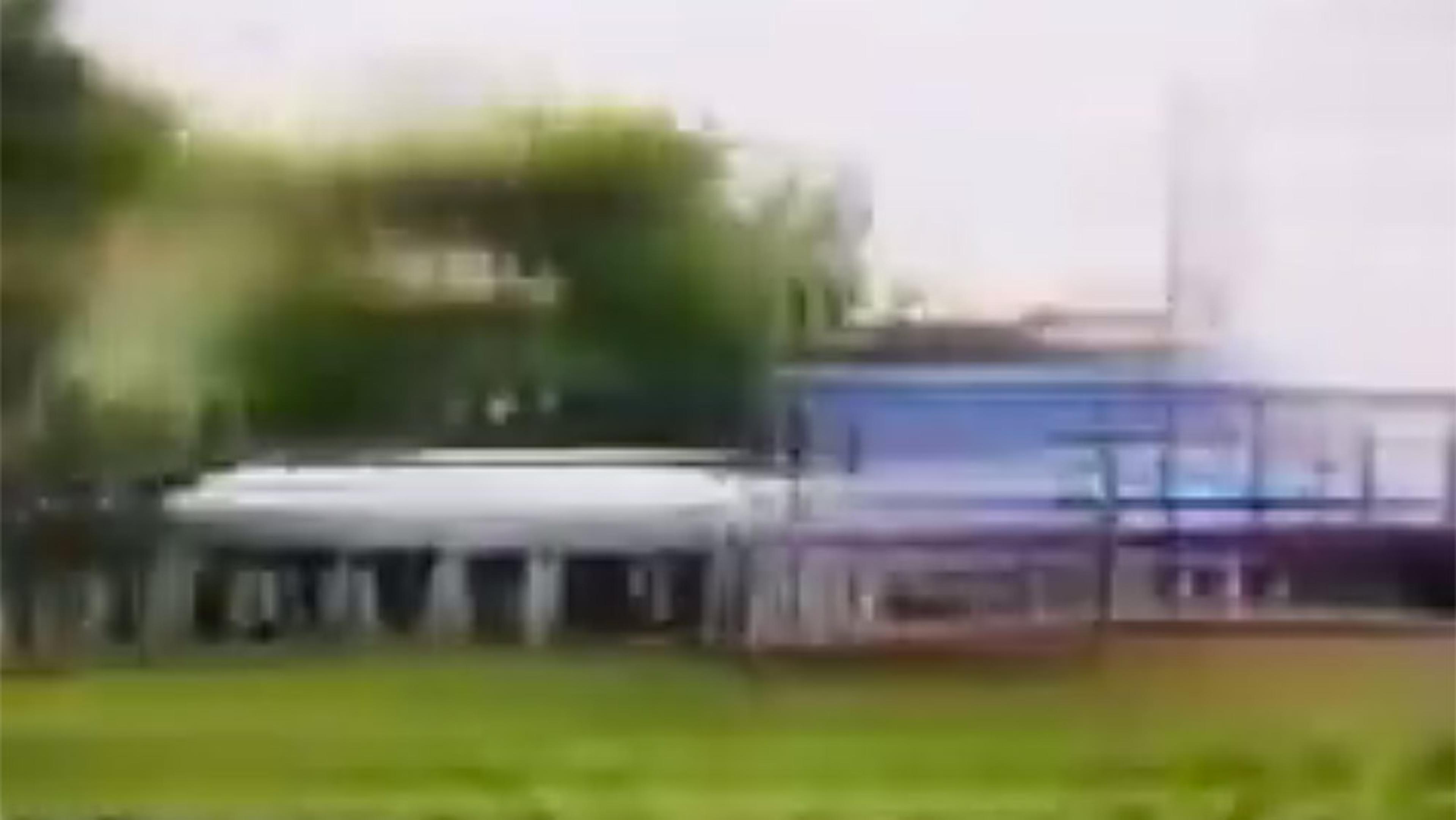 Blurry photo of a rectangular structure with a white canopy and columns, set in a grassy area with trees in the background.