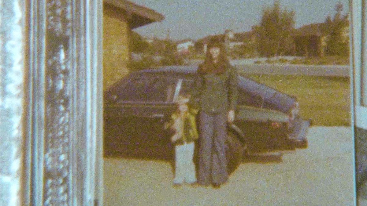 Vintage photo of a women and child standing in front of a dark car parked in a driveway, with a suburban background.