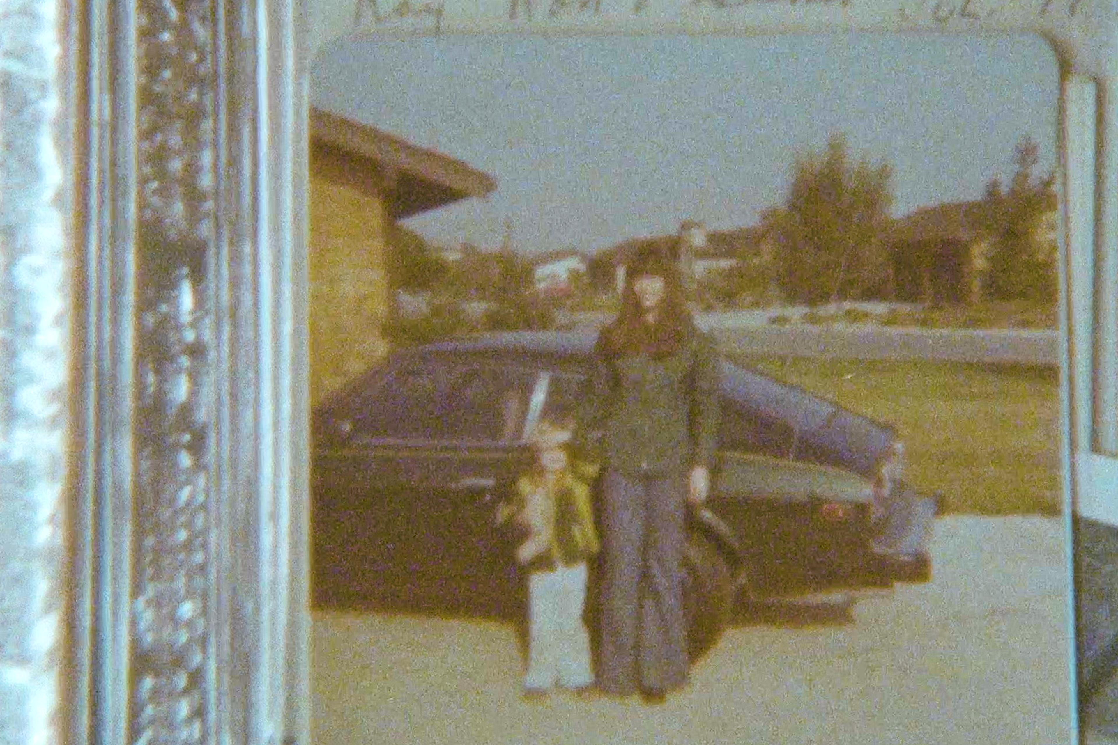 Vintage photo of a women and child standing in front of a dark car parked in a driveway, with a suburban background.