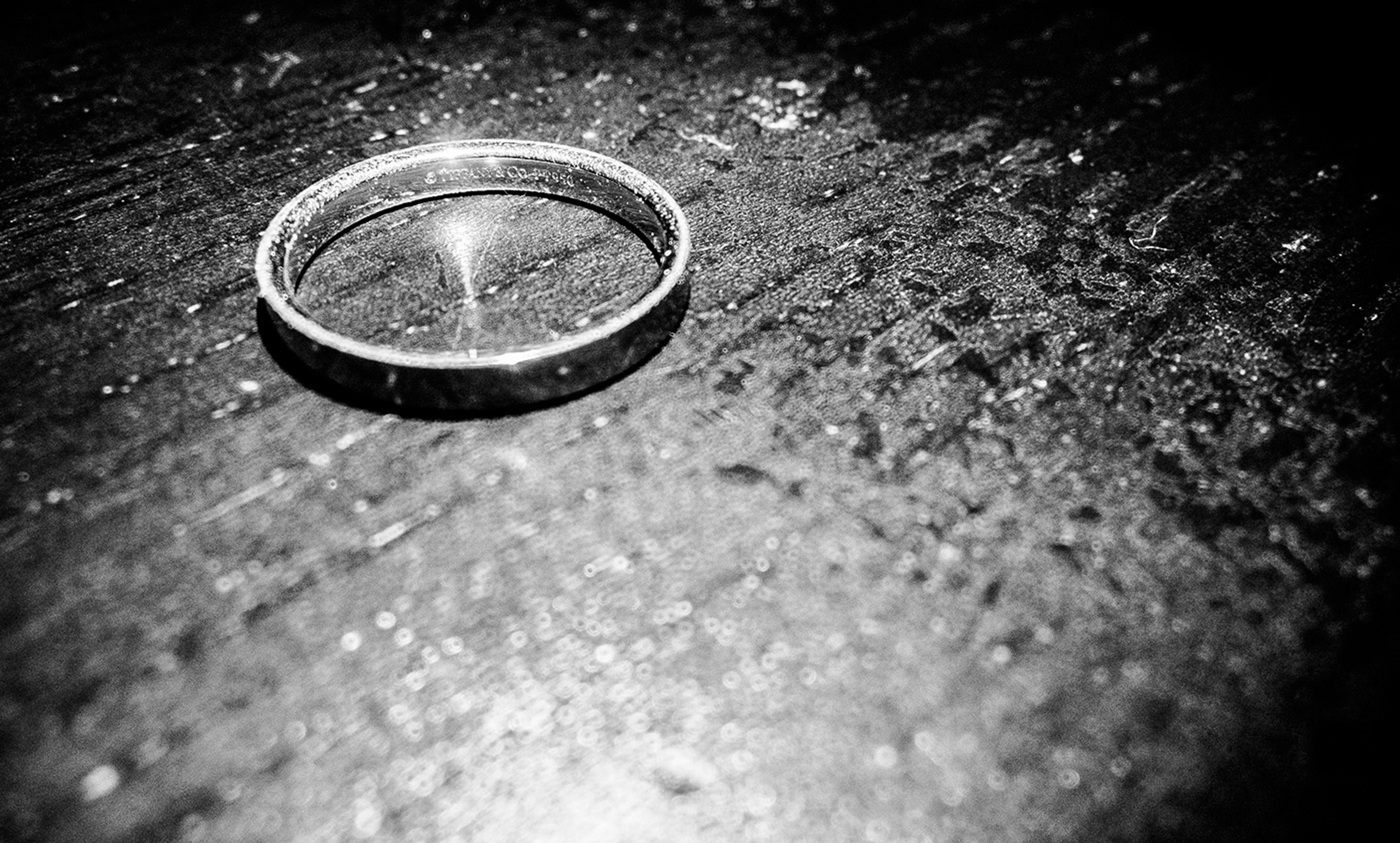 Black and white photo of a single ring on a textured dark surface, illuminated to create a dramatic shadow and reflection effects.