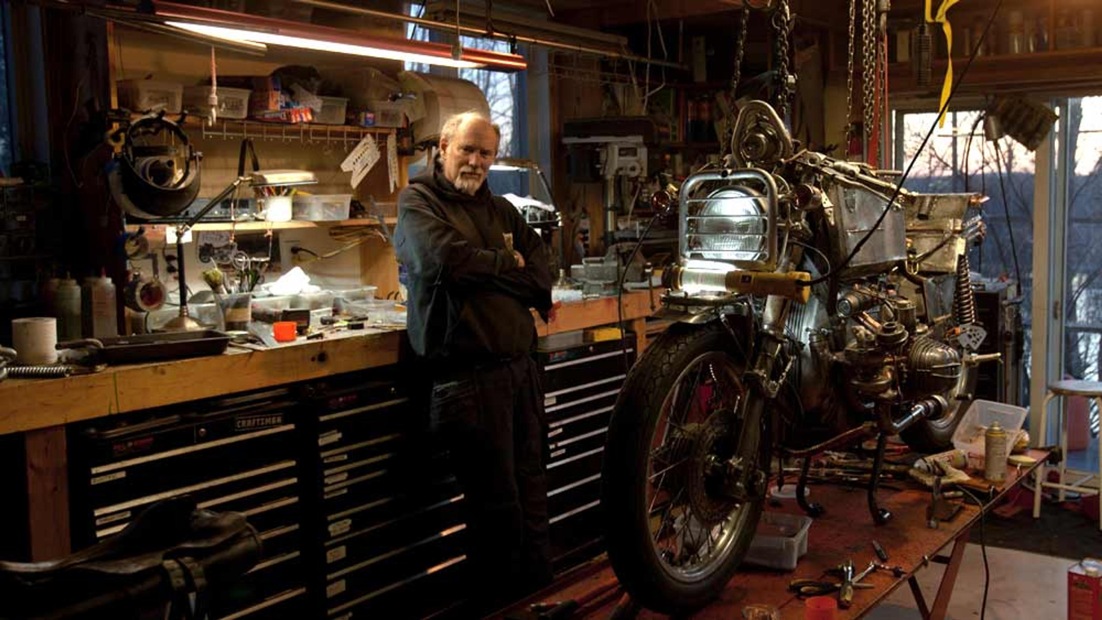 A man in a workshop standing next to a workbench and a motorcycle being repaired, with tools and parts around.