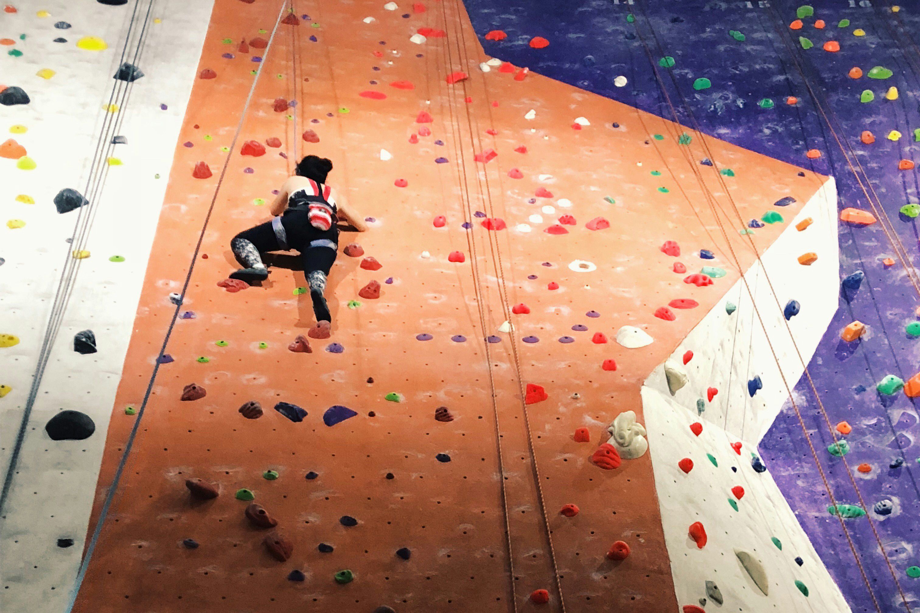 A person climbing a colourful indoor rock wall with various holds and ropes visible.
