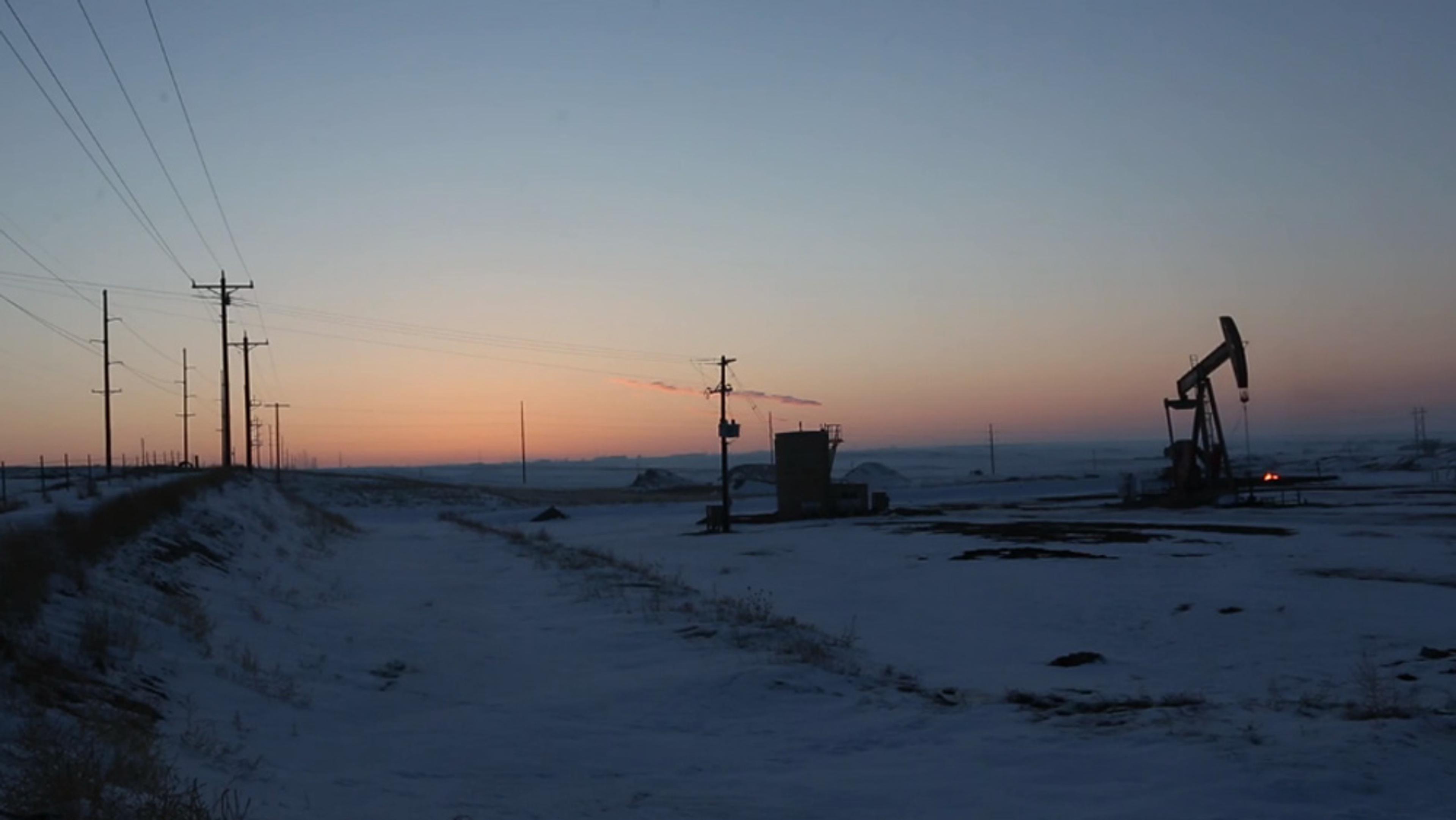 An oil pump in a snowy landscape at dusk, with power lines and poles stretching into the distance under a dimly lit sky.