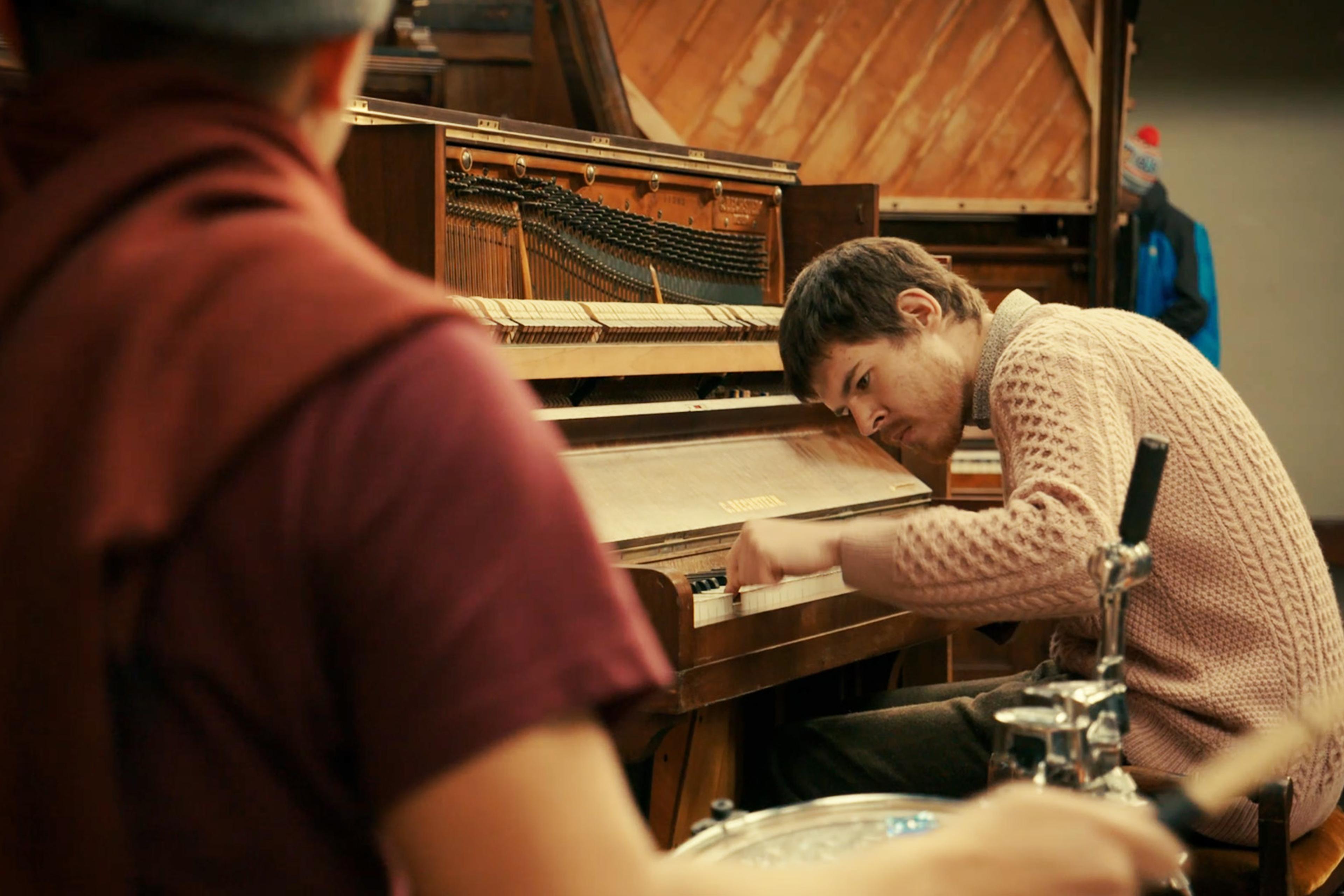 Man in a pink jumper intensely plays a piano in a cosy room with a wooden interior, while another person, blurred in the foreground, plays drums.