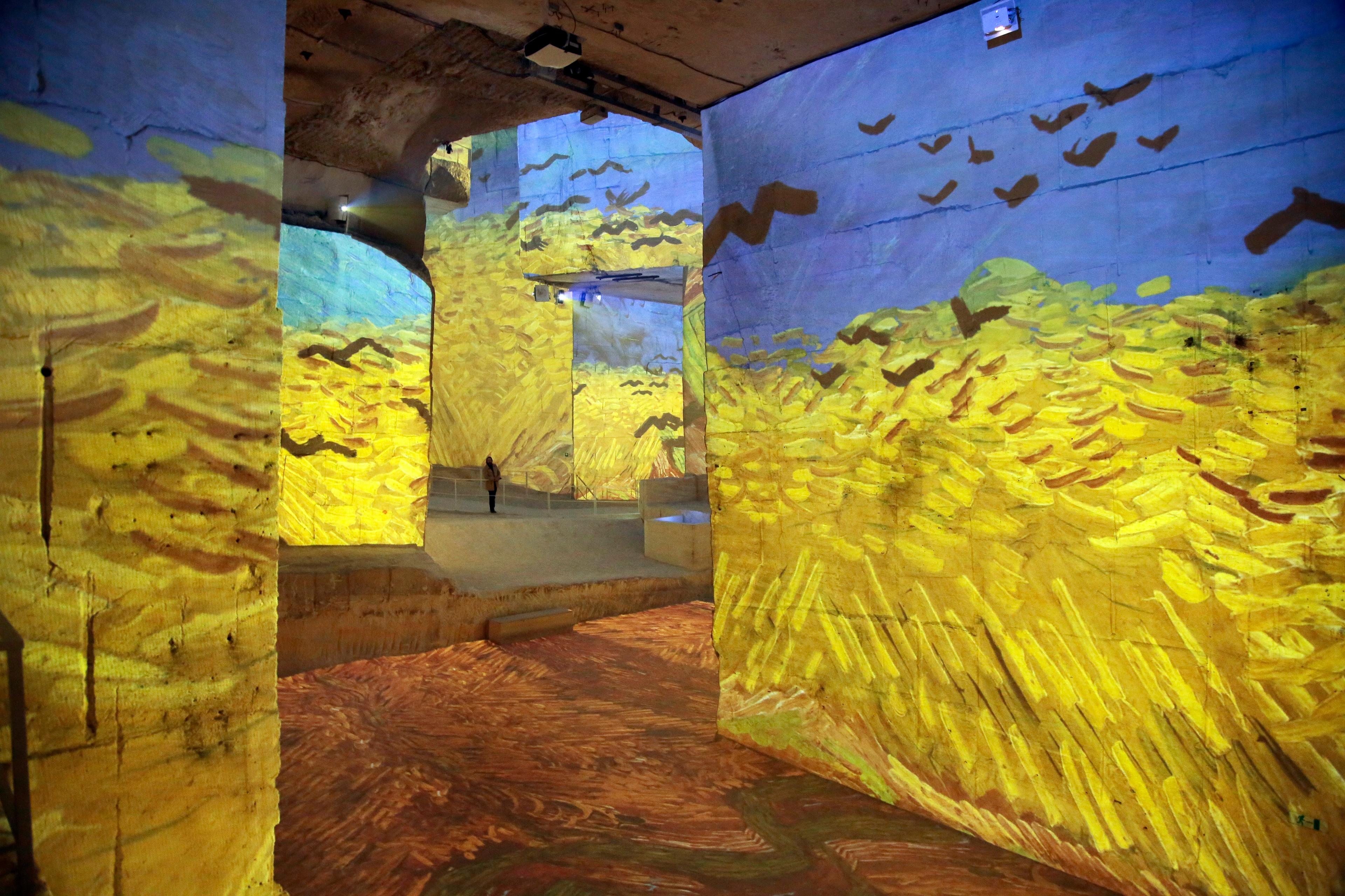 Gallery walls with a projection of a vibrant wheat field painting and a person observing the immersive scene.