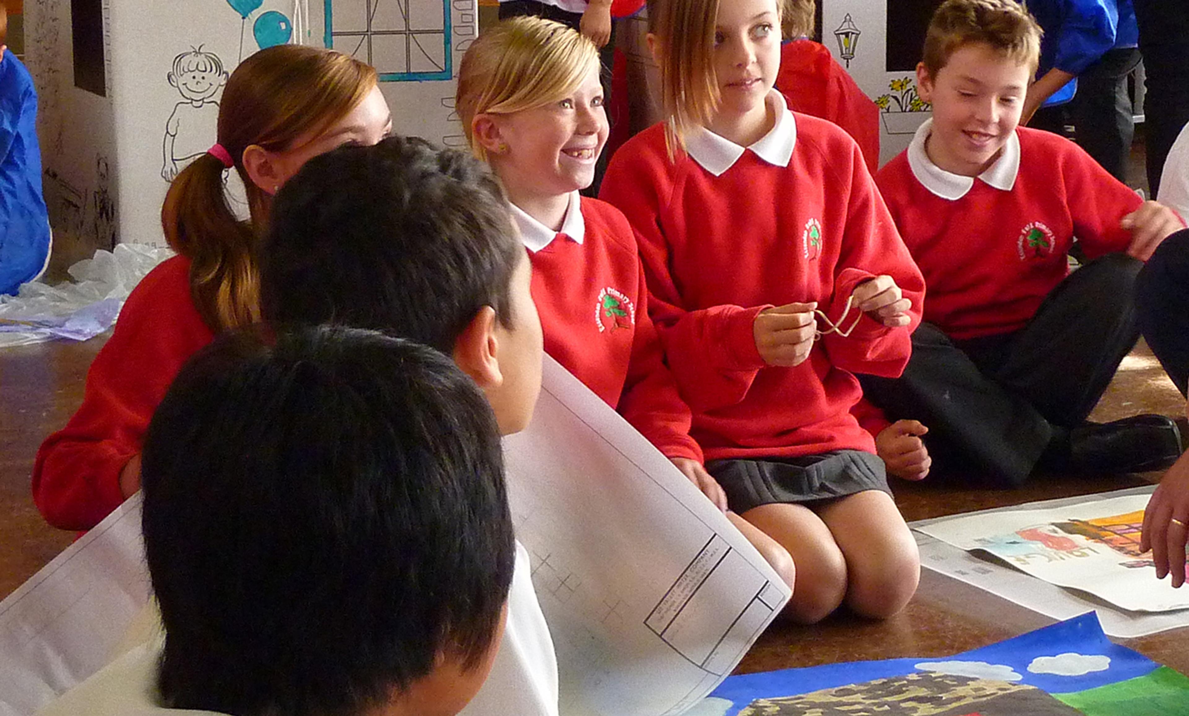 Schoolchildren in red jumpers sitting on the floor, holding drawings and papers, engaging in a group activity.
