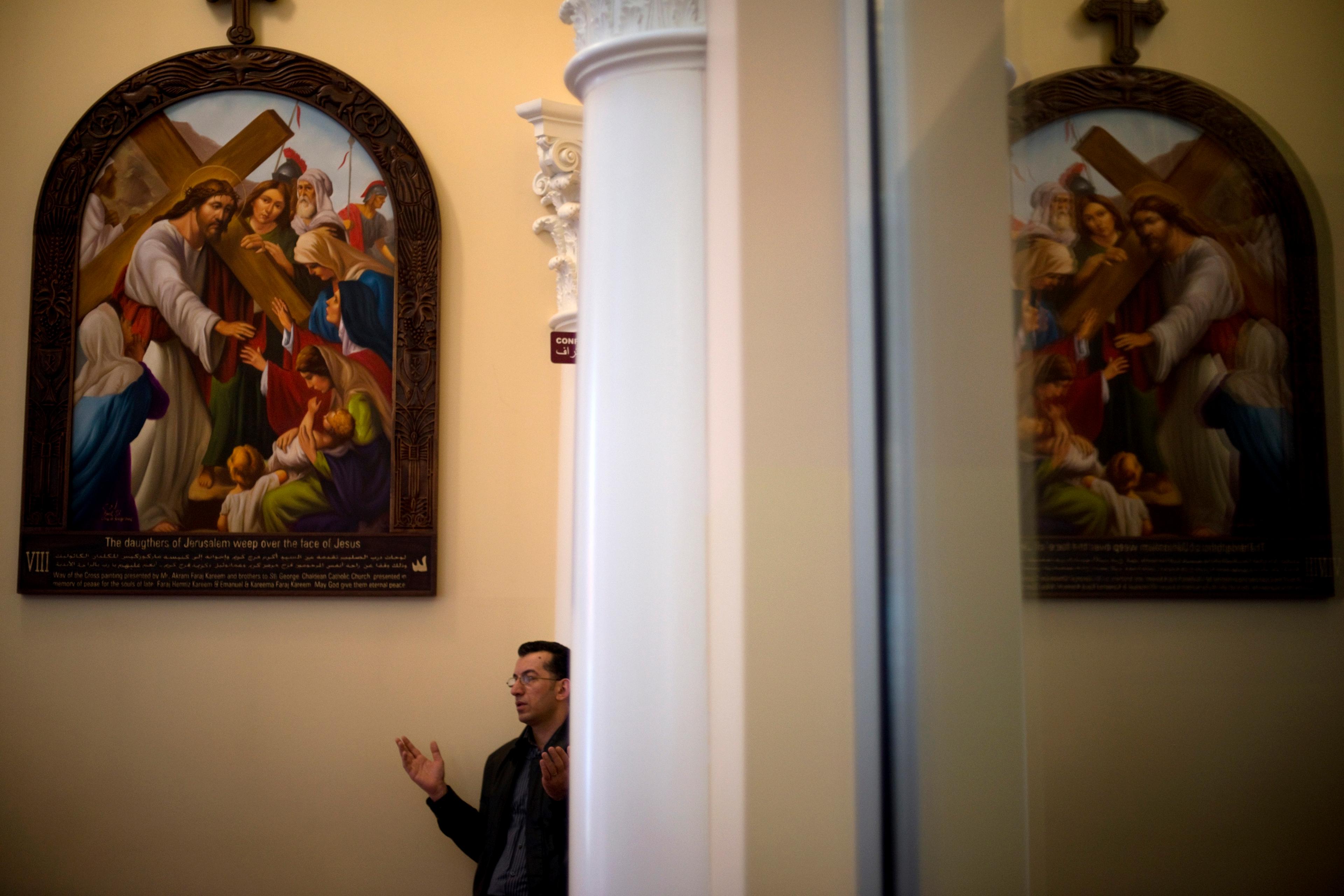 Photo of a man praying next to a large religious painting in a church with ornate columns in the foreground.