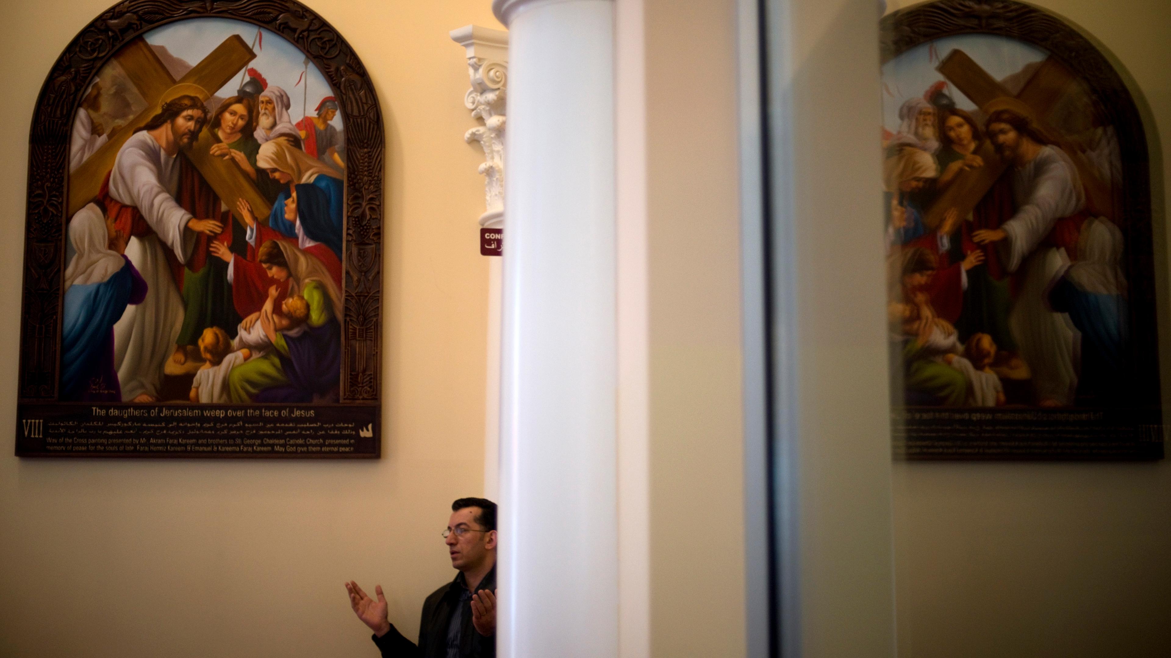 Photo of a man praying next to a large religious painting in a church with ornate columns in the foreground.