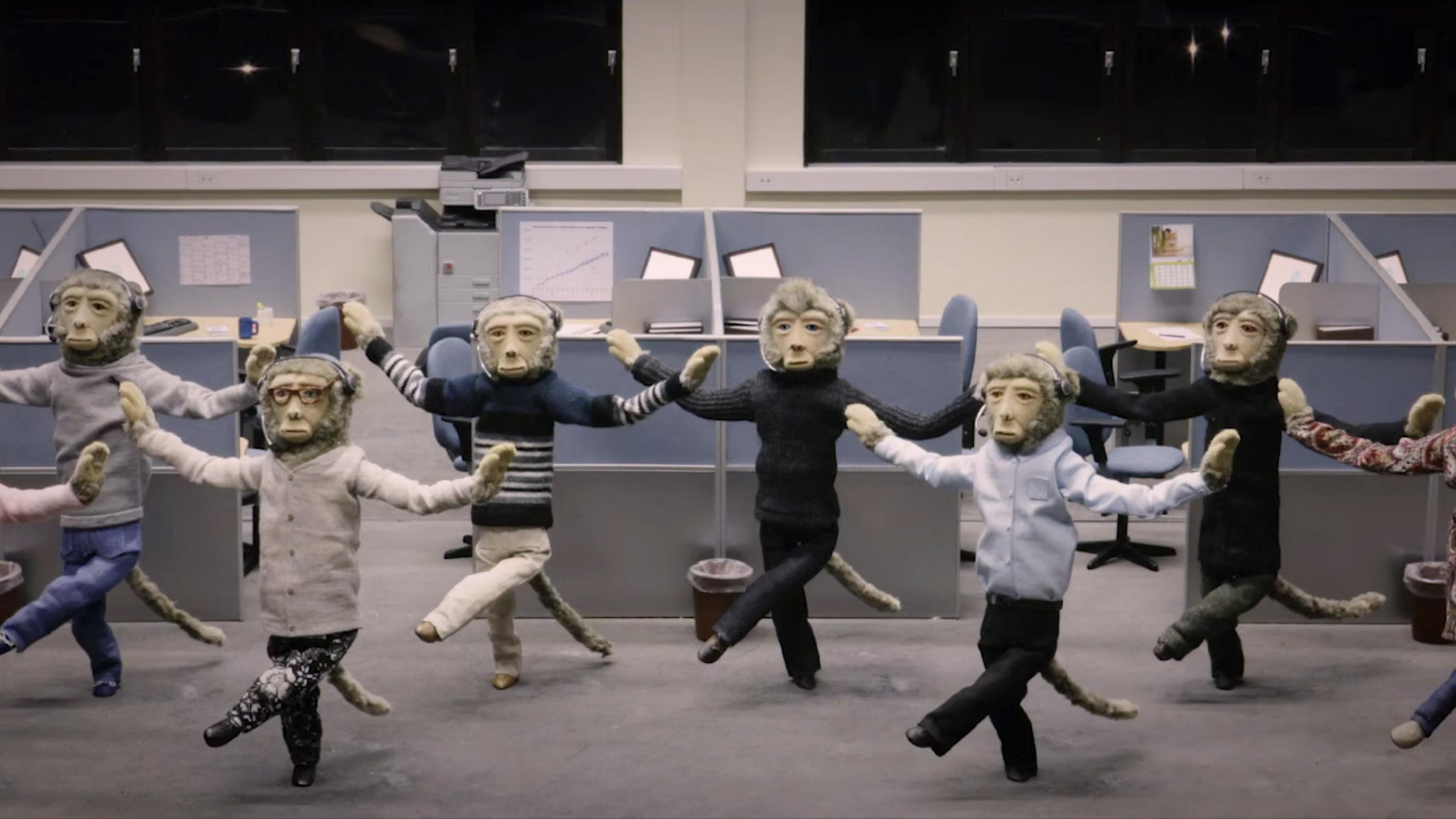 Puppets with monkey faces in office attire, performing a synchronised dance in a cubicle-filled office setting, with computers and office supplies visible.