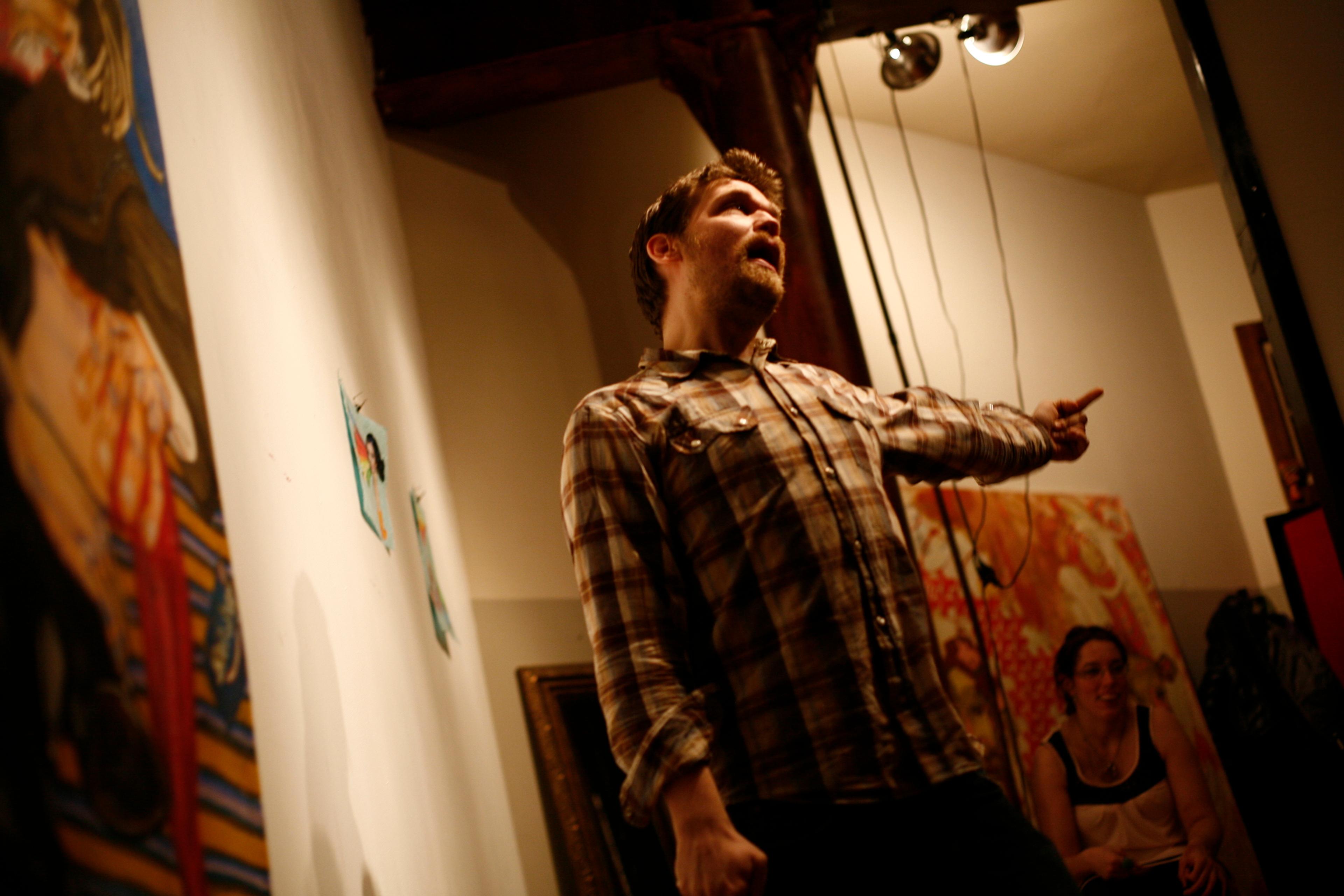 Photo of a man in a plaid shirt enthusiastically gesturing in an art-filled room with a seated woman in the background.