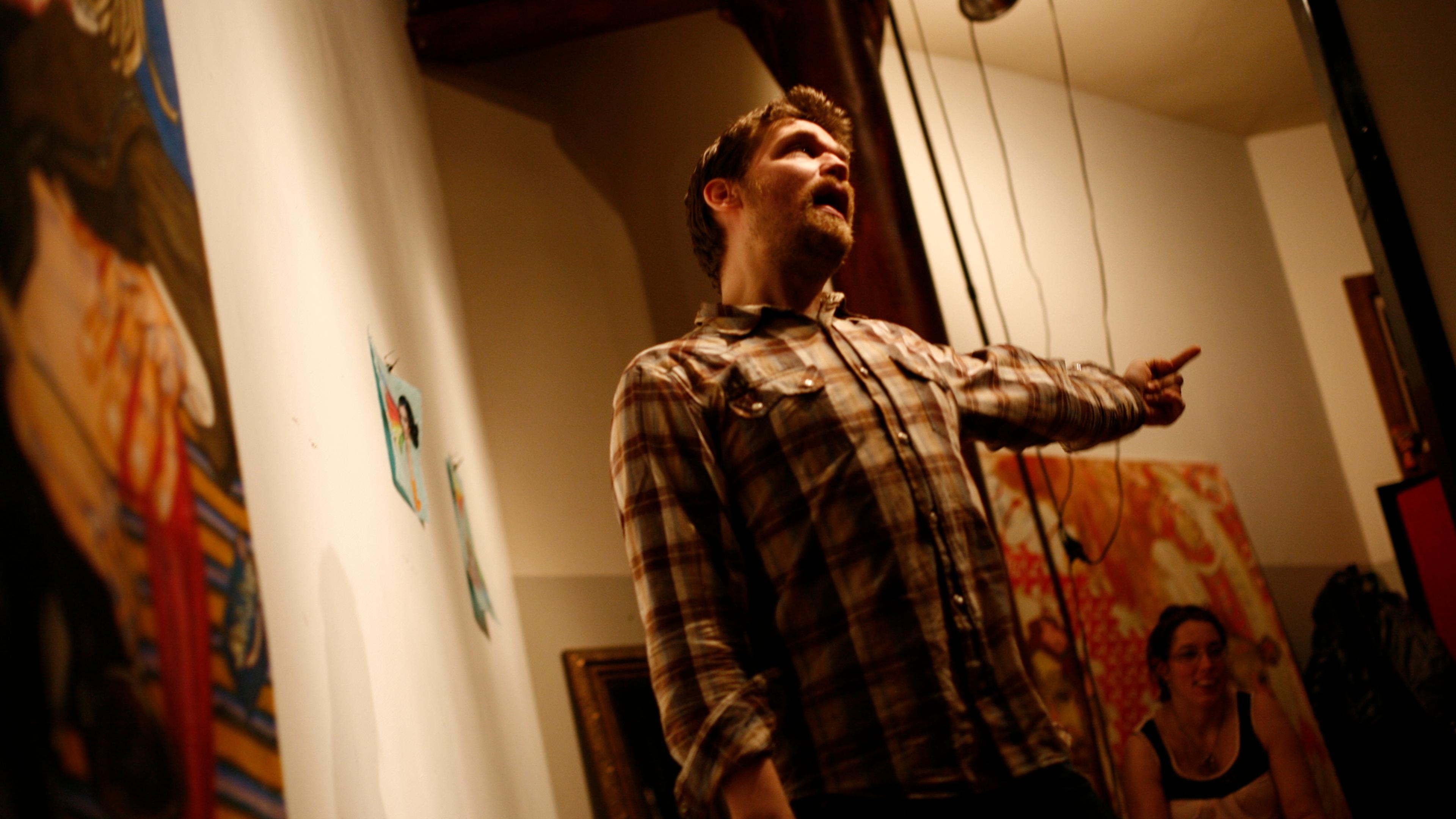 Photo of a man in a plaid shirt enthusiastically gesturing in an art-filled room with a seated woman in the background.