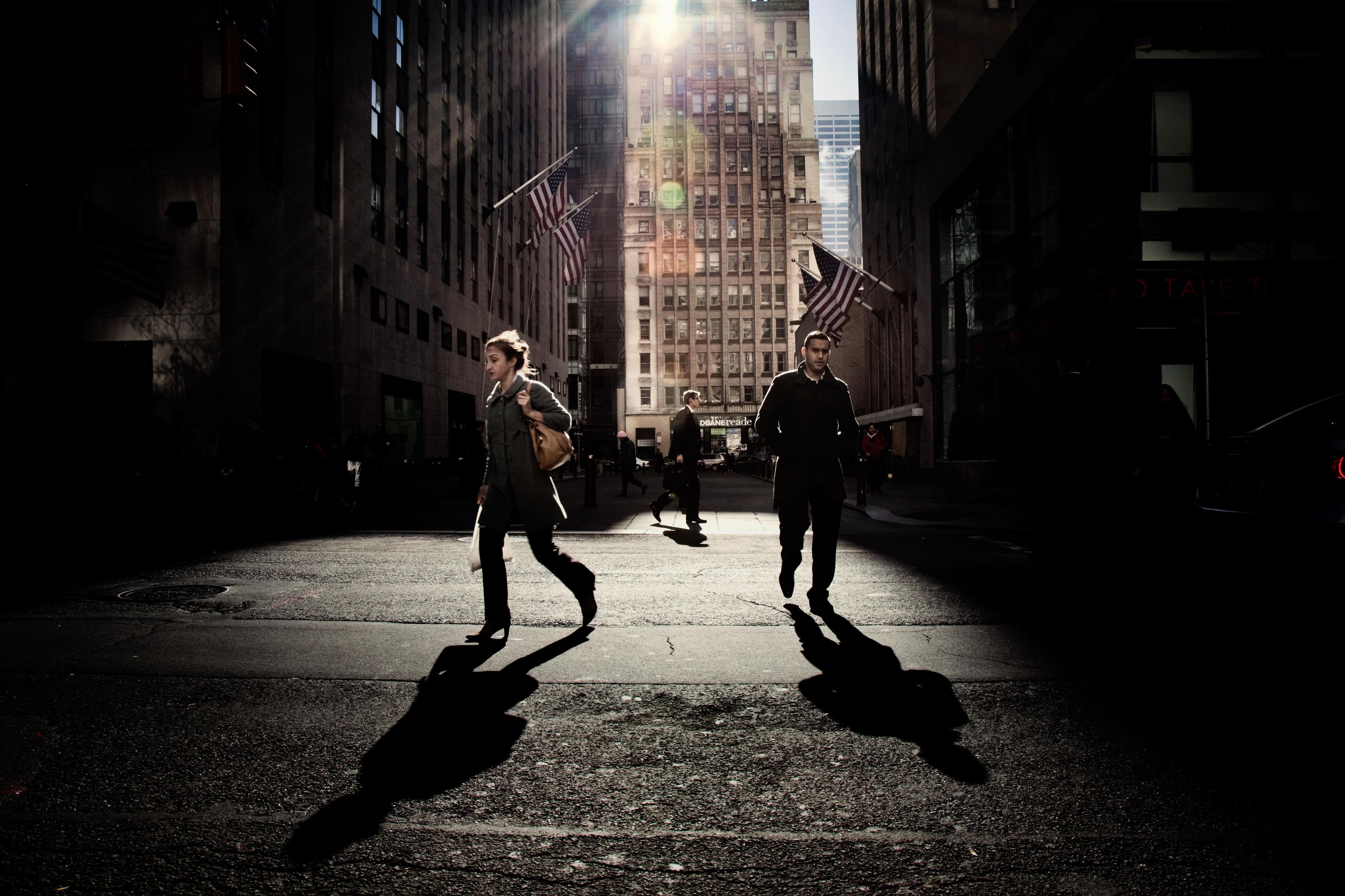 Photo of people crossing a street in a city. Sunlight casts long shadows. Skyscrapers and flags are in the background.