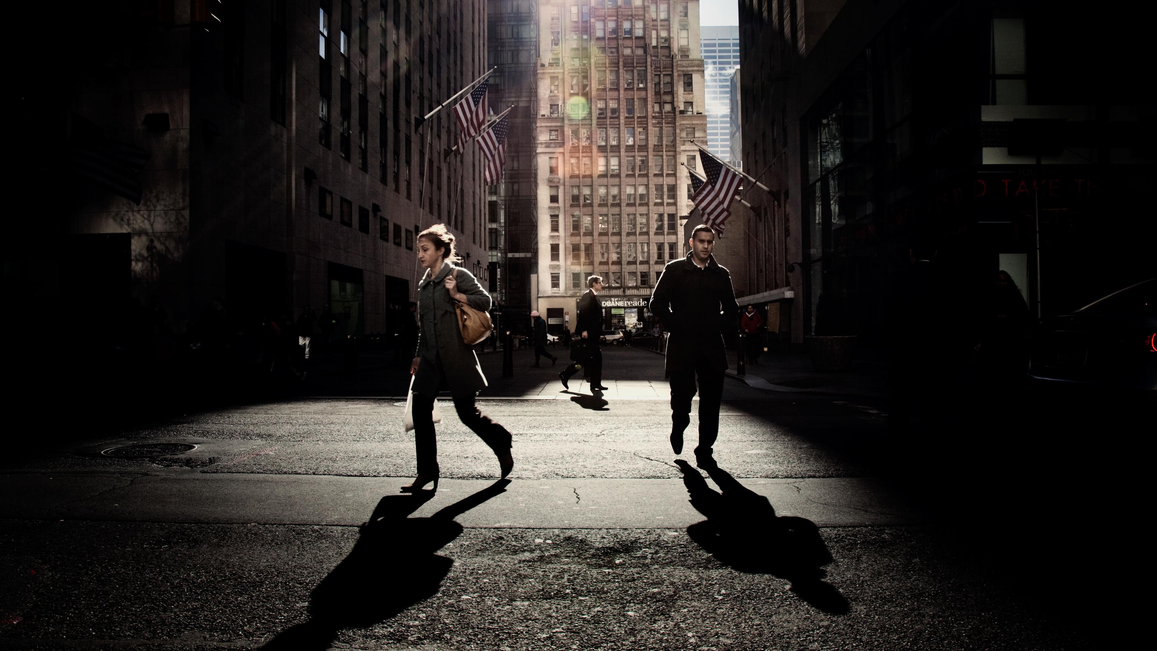 Photo of people crossing a street in a city. Sunlight casts long shadows. Skyscrapers and flags are in the background.