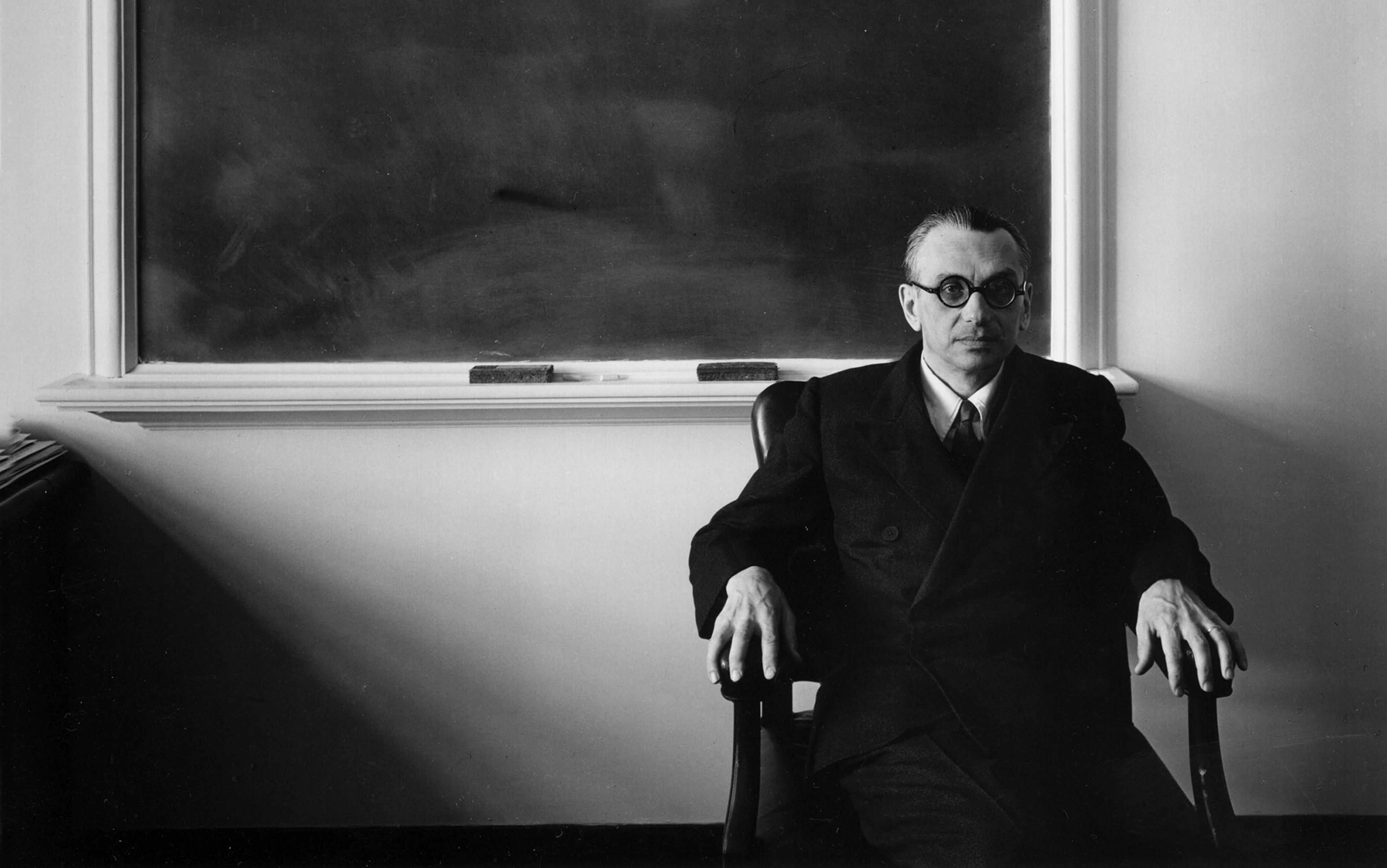 Black-and white photo of a man wearing glasses and a suit sitting in a chair in front of a blackboard in a classroom.