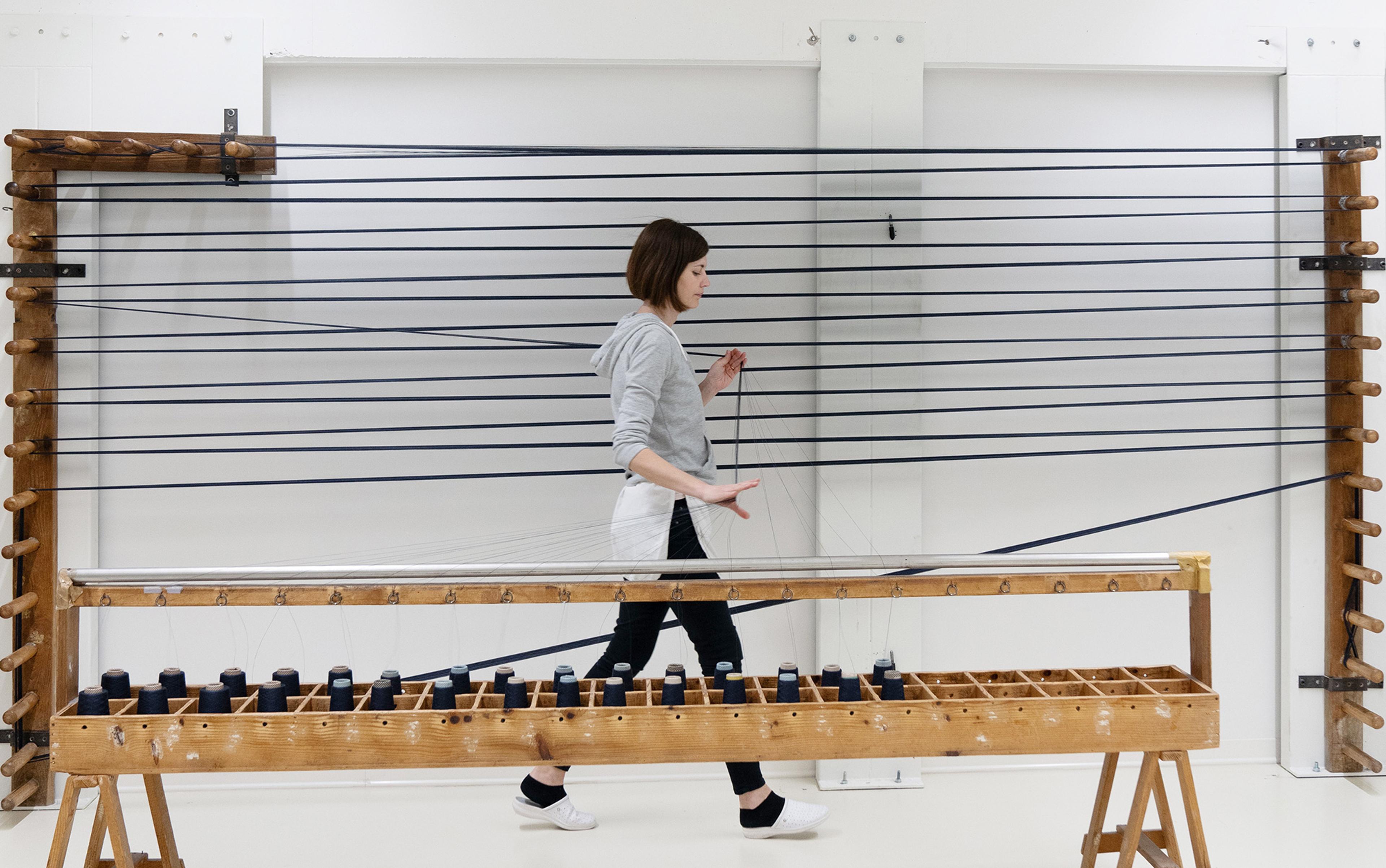 A woman in casual attire walking by a large weaving loom with black threads in a bright, clean room