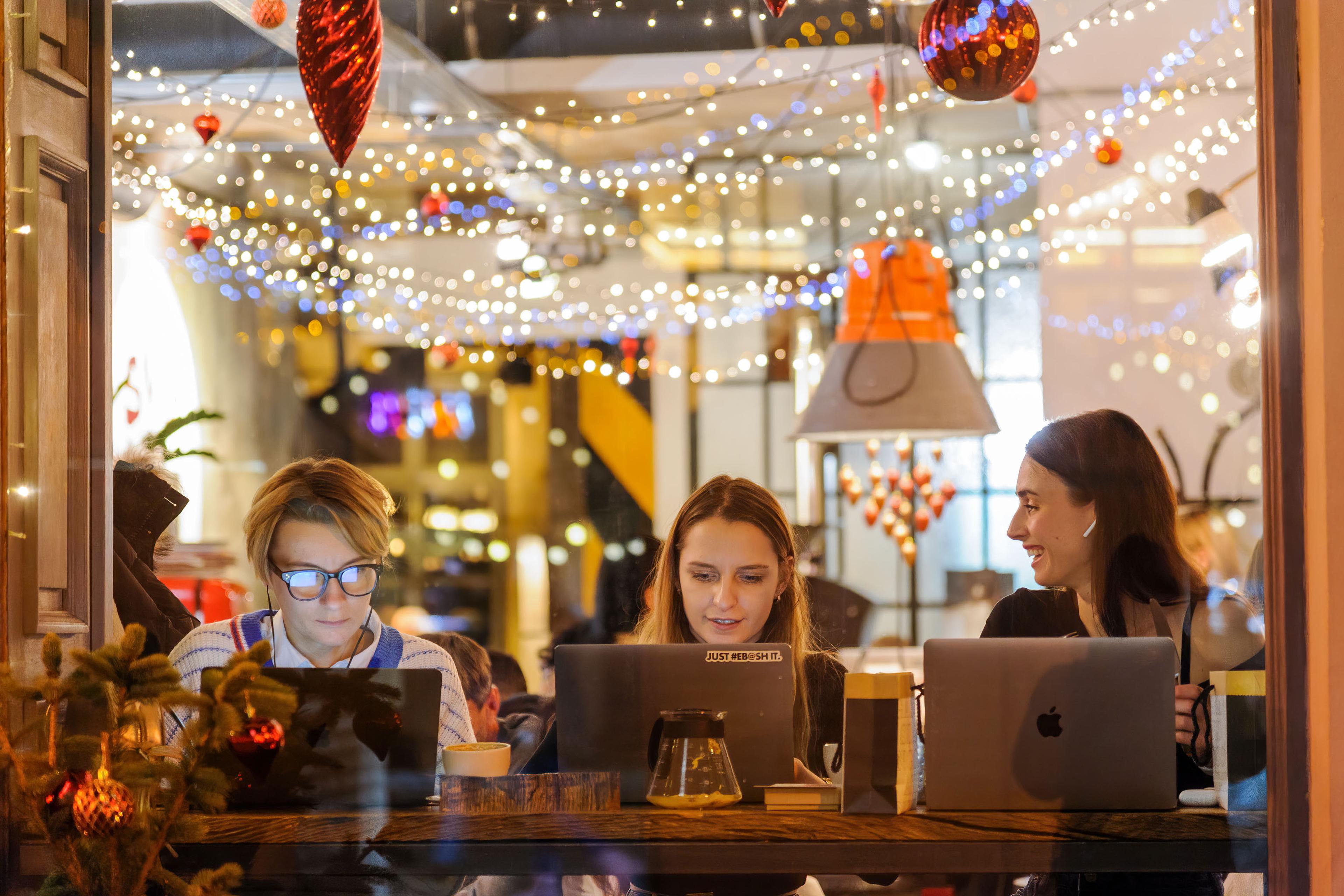 Photo of three women working on laptops in a café with festive lights and decorations hanging from the ceiling.