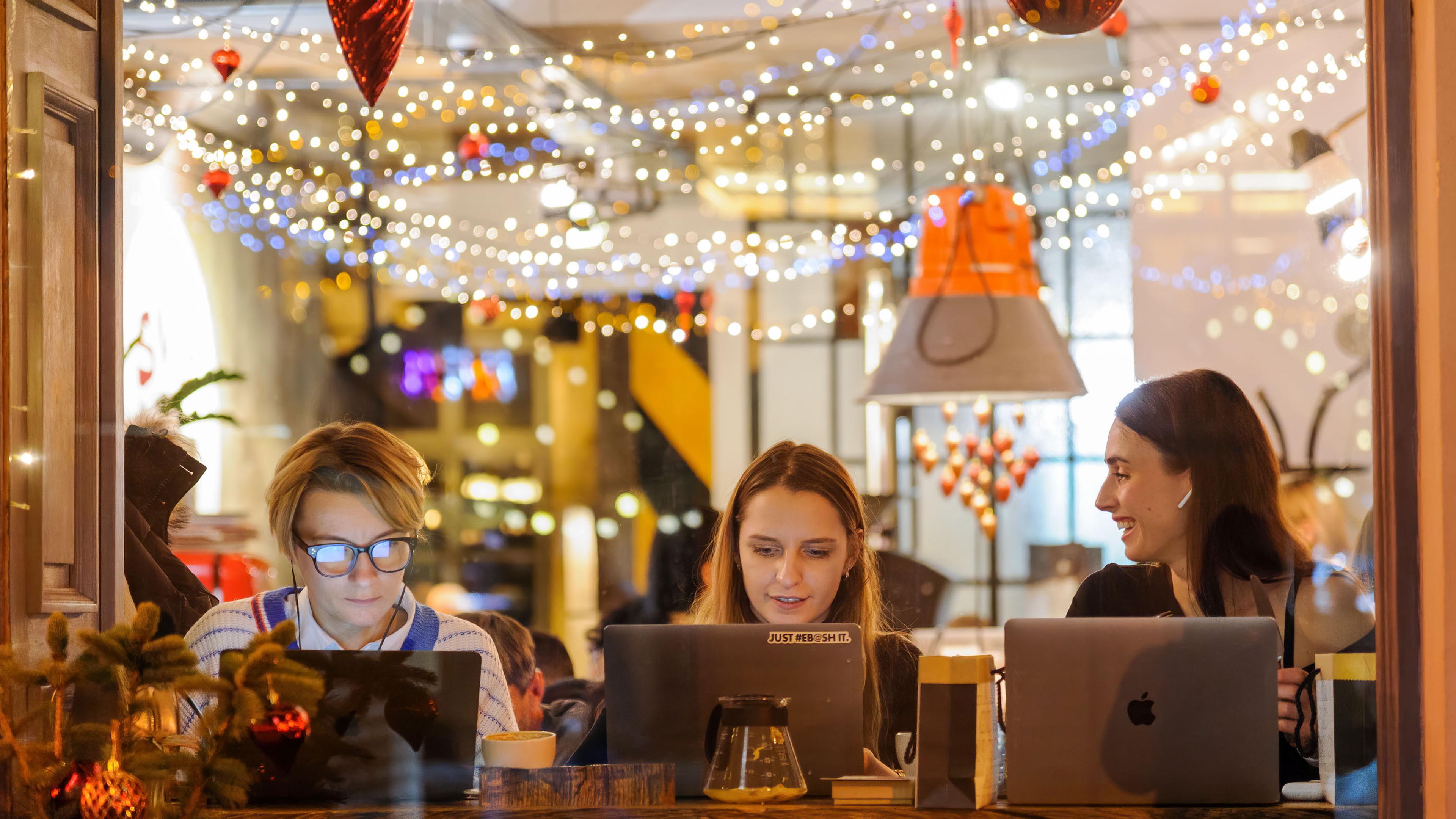 Photo of three women working on laptops in a café with festive lights and decorations hanging from the ceiling.