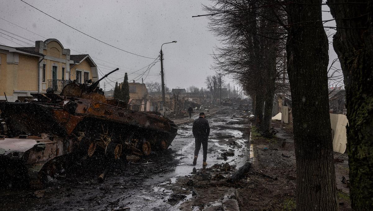 A man walks beneath overcast skies down a wet and rutted road past a burned out tank in war-ravaged Ukraine