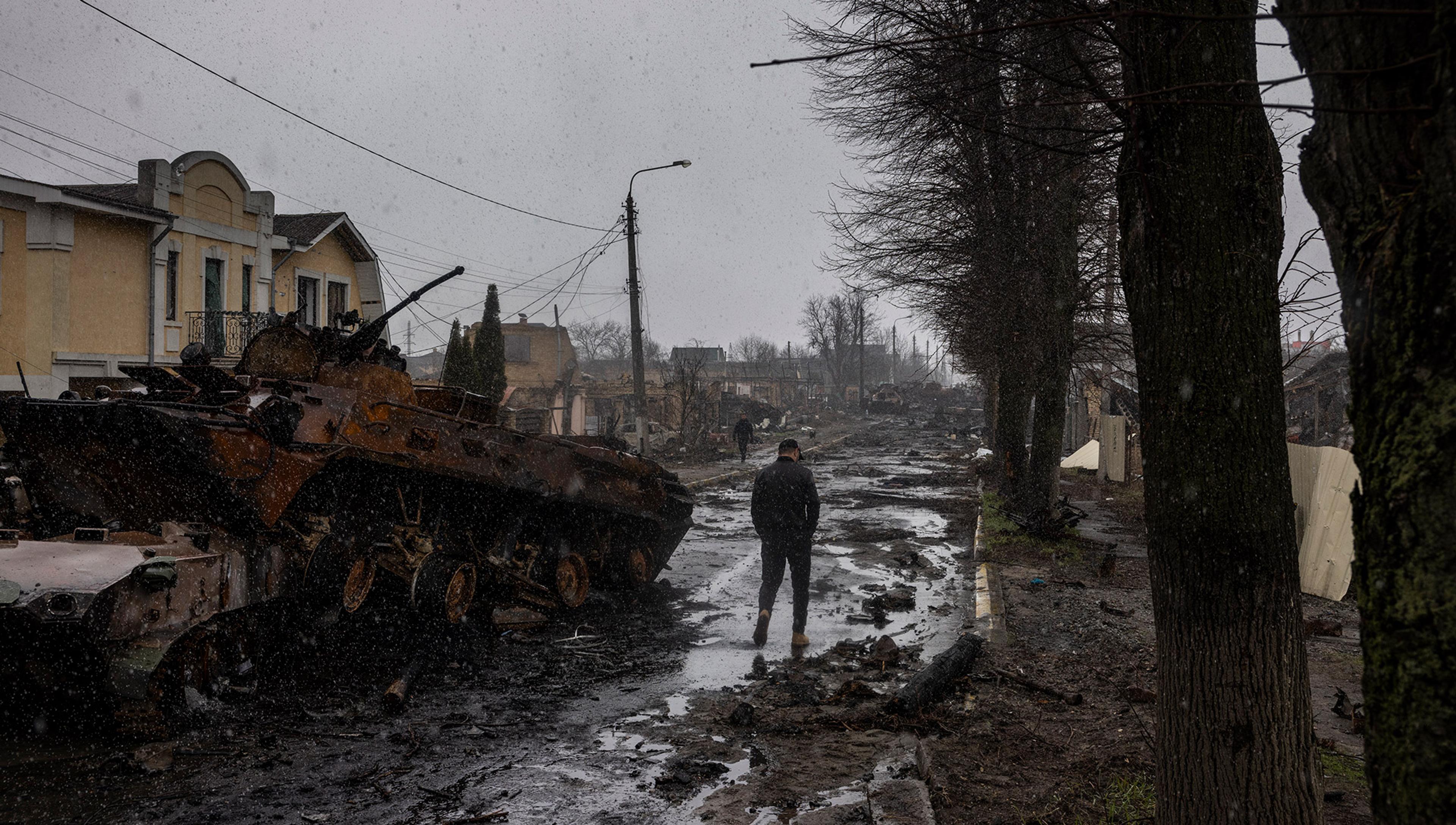 A man walks beneath overcast skies down a wet and rutted road past a burned out tank in war-ravaged Ukraine