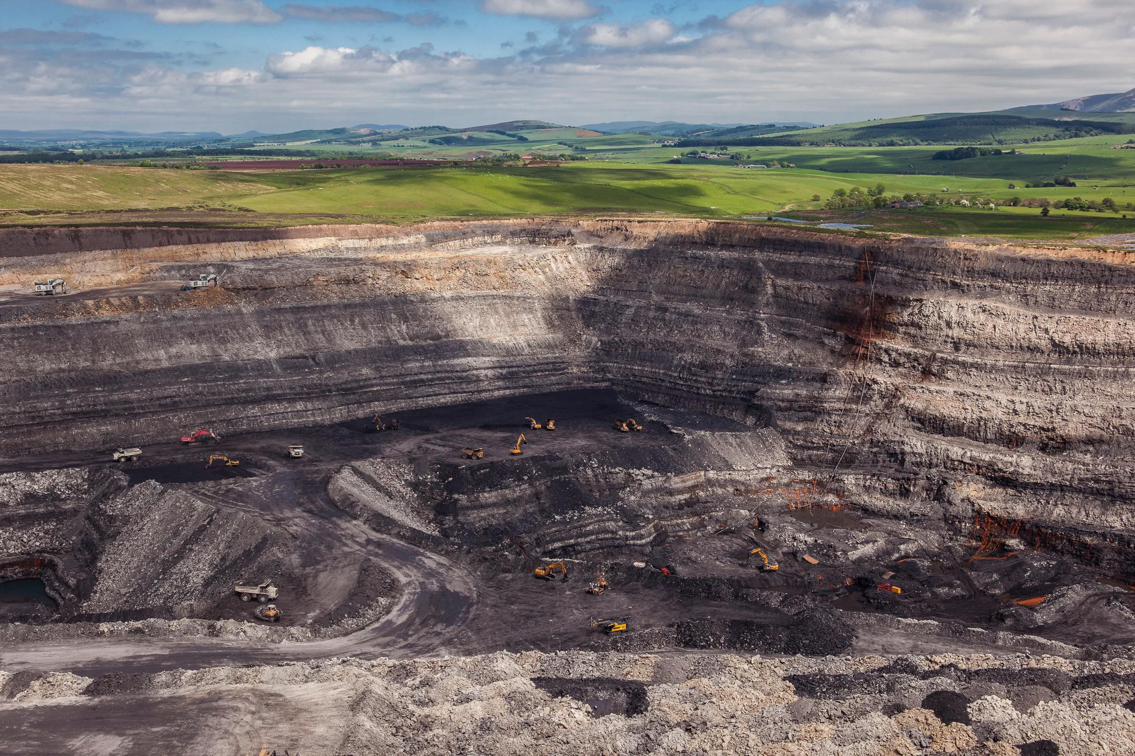 A vast open-pit coal mine with excavation machinery surrounded by green hills.