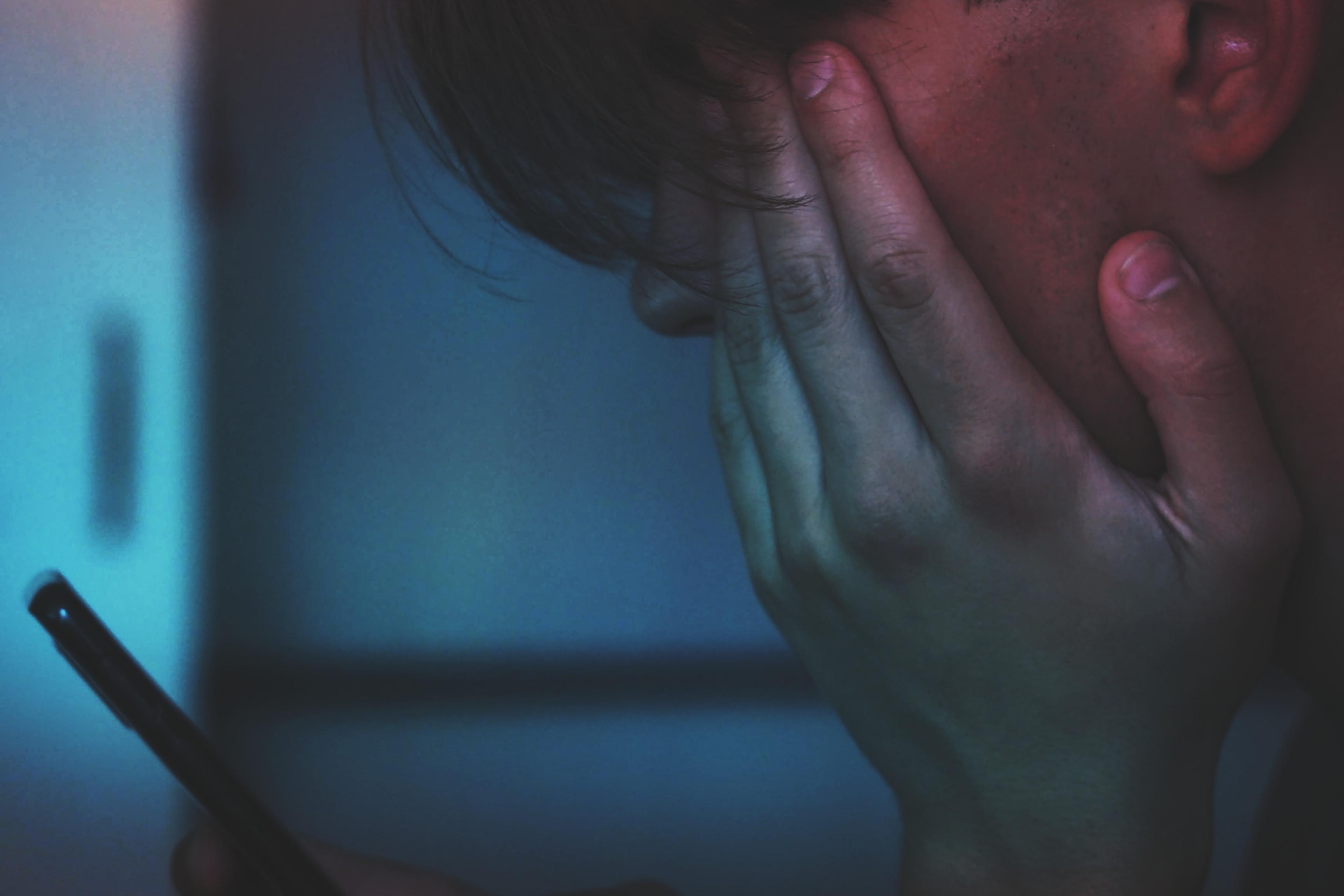 Photo of a person holding a smartphone, face rested on their hand, illuminated by blue and red light.