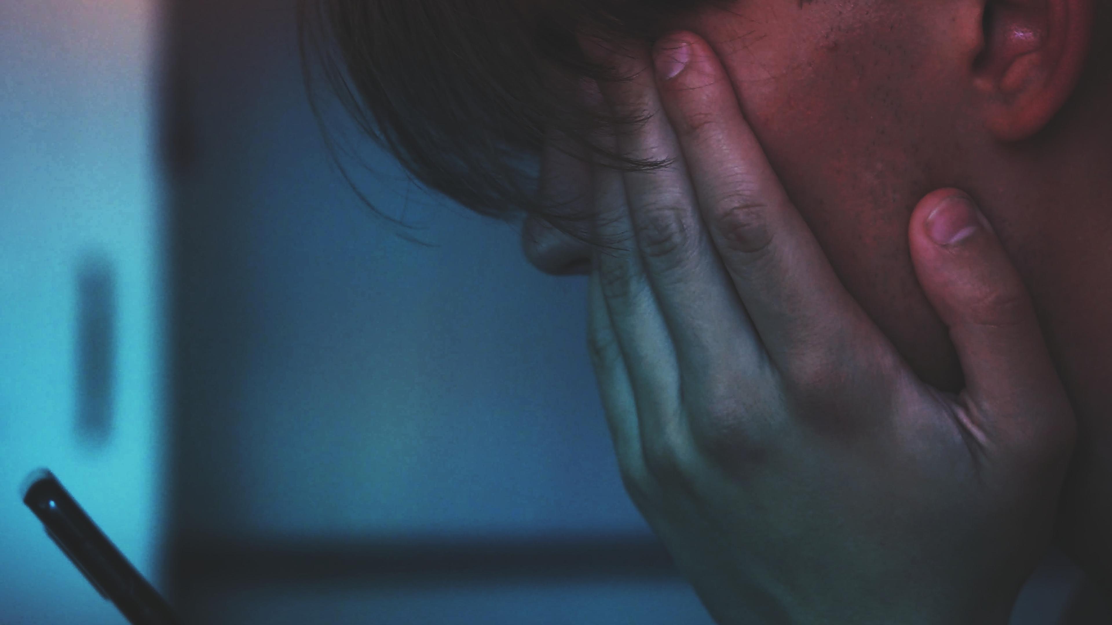 Photo of a person holding a smartphone, face rested on their hand, illuminated by blue and red light.