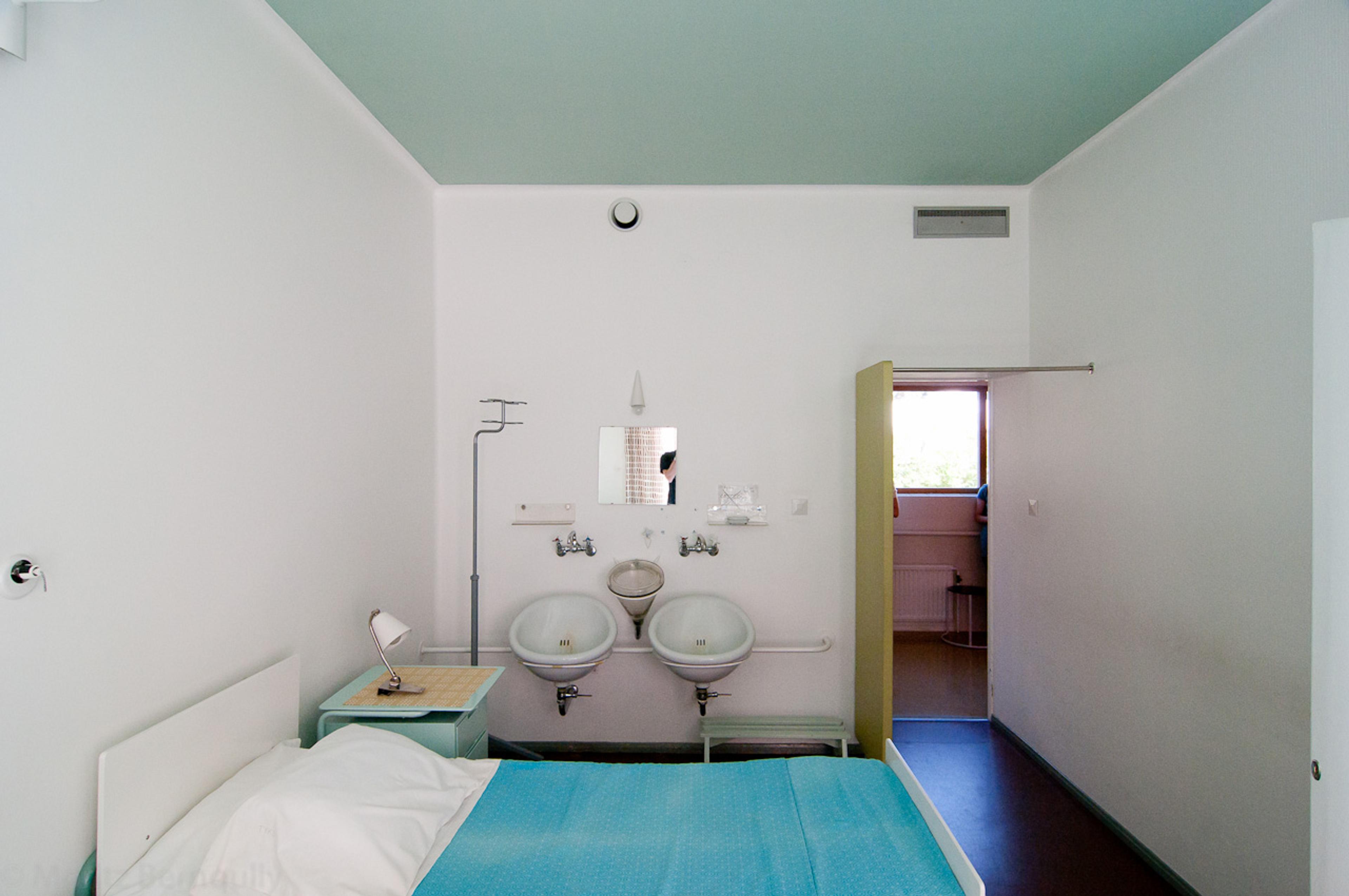 A hospital room with a single bed, two washbasins, a nightstand, and an open door, all under pastel green and white colours.