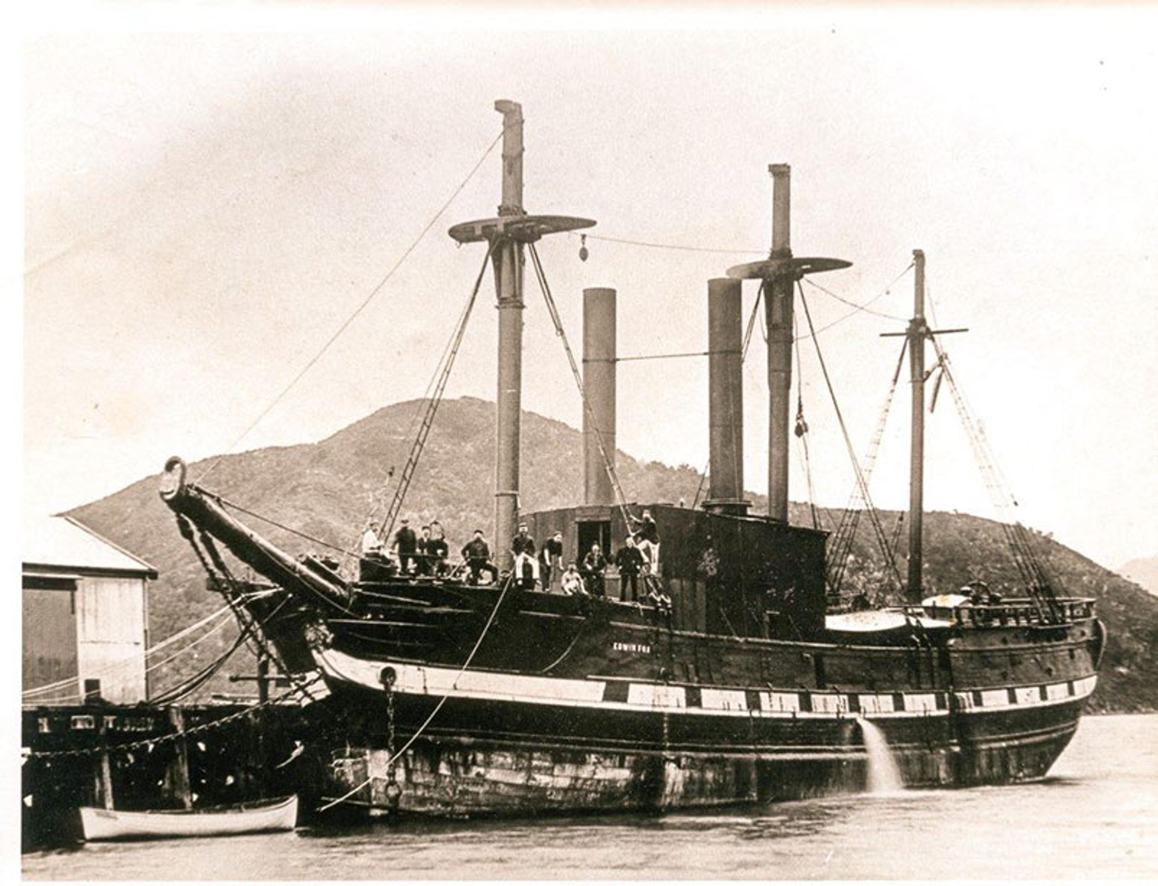 Historic photo of a large ship with two masts docked by a mountainous backdrop, with people visible on deck and beside the ship