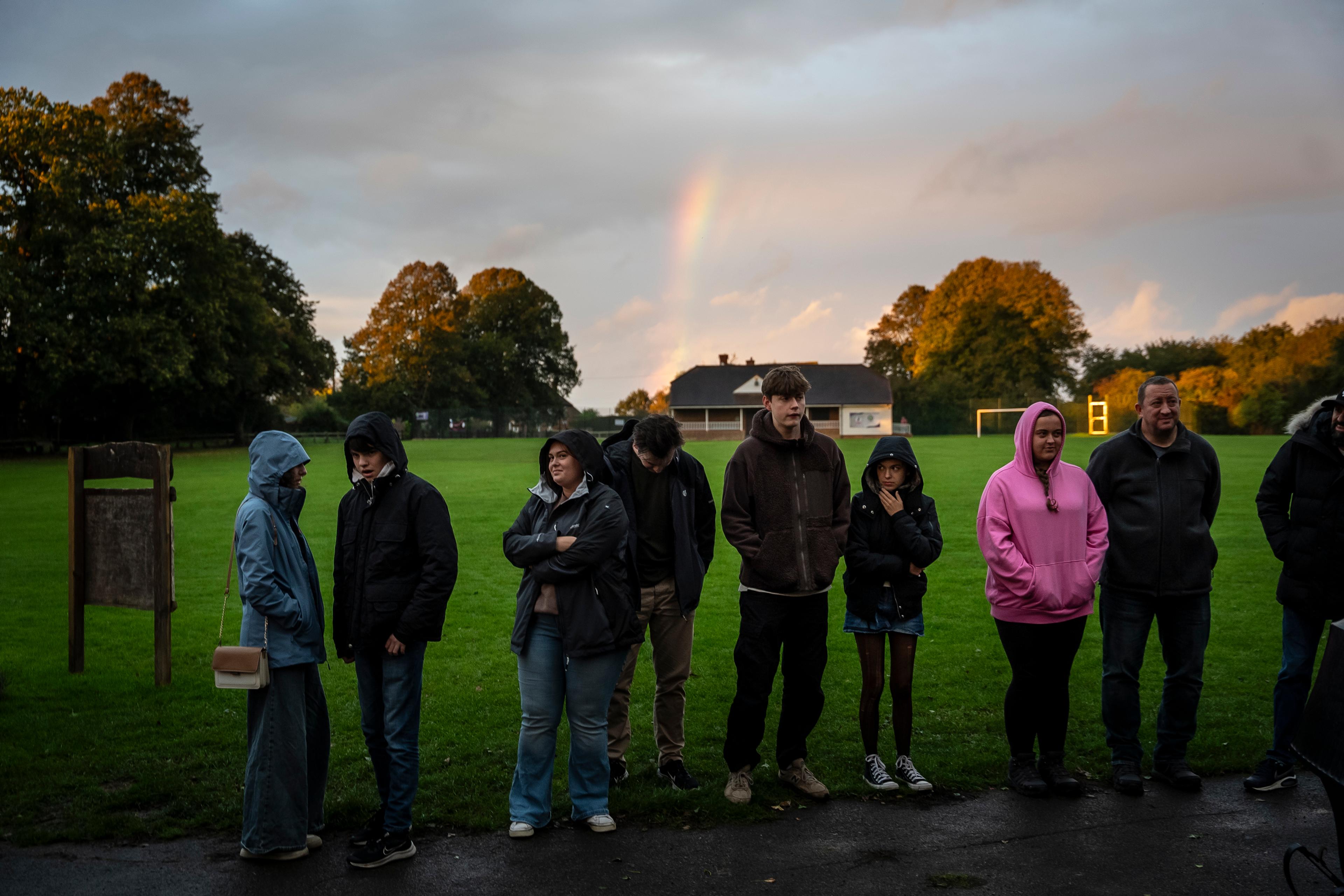 People in hooded jackets standing in a park with grass and trees, a rainbow in the sky and a building in the background.