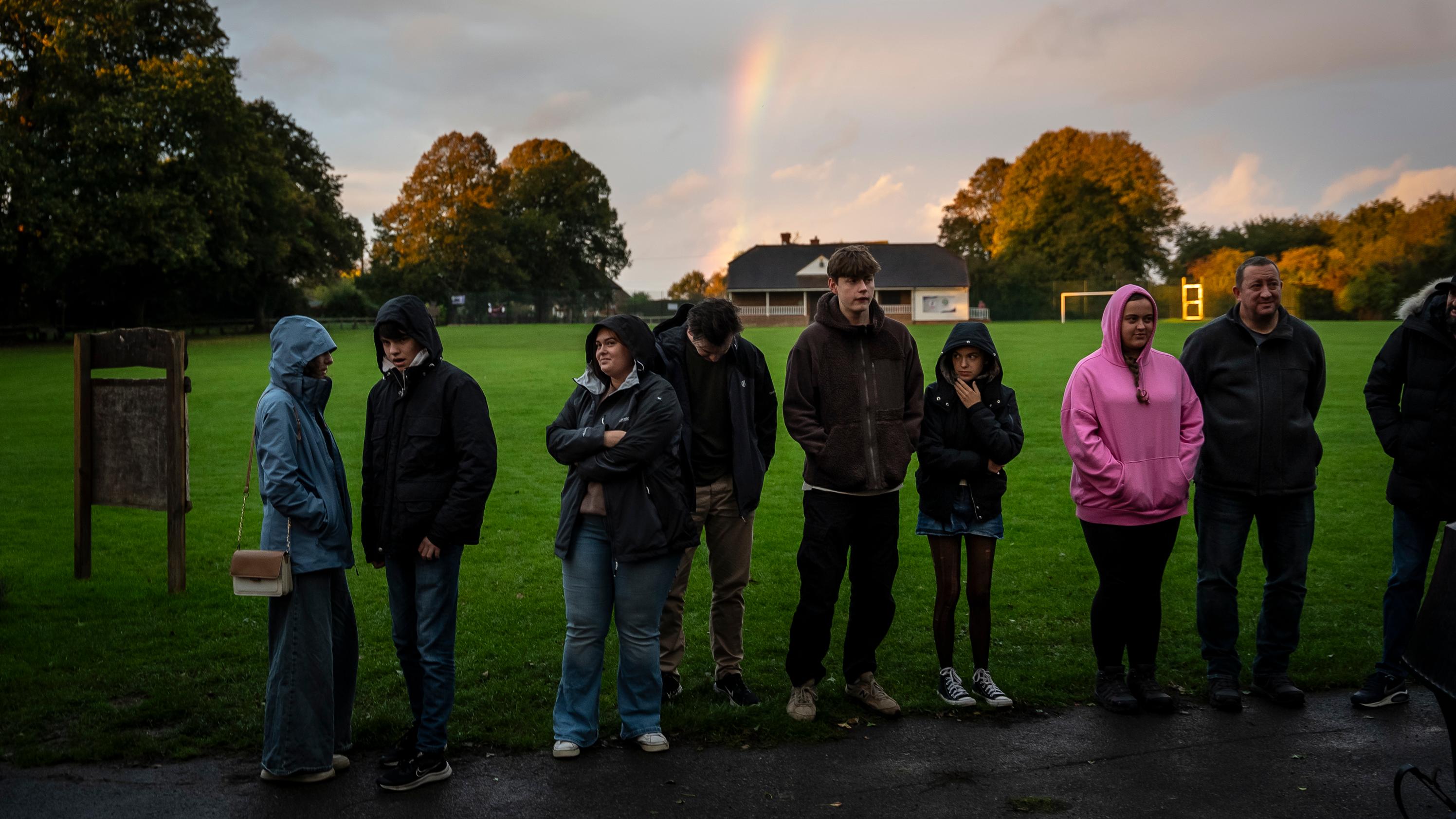 People in hooded jackets standing in a park with grass and trees, a rainbow in the sky and a building in the background.
