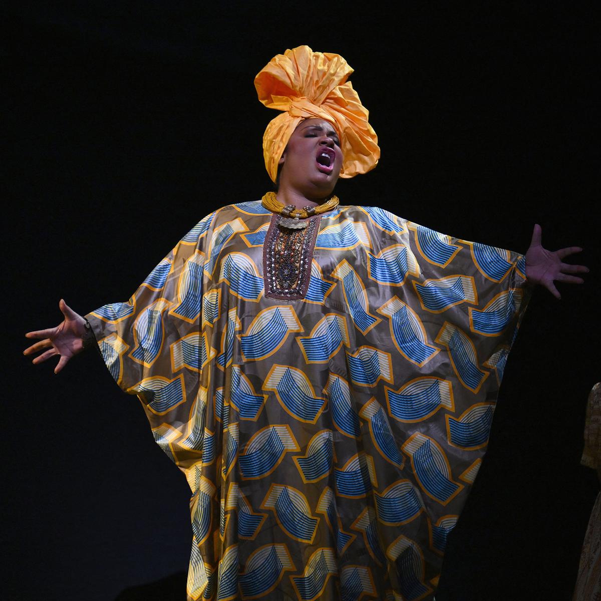 Photo of a person singing dramatically in colourful patterned attire and an orange headwrap on a dark stage.