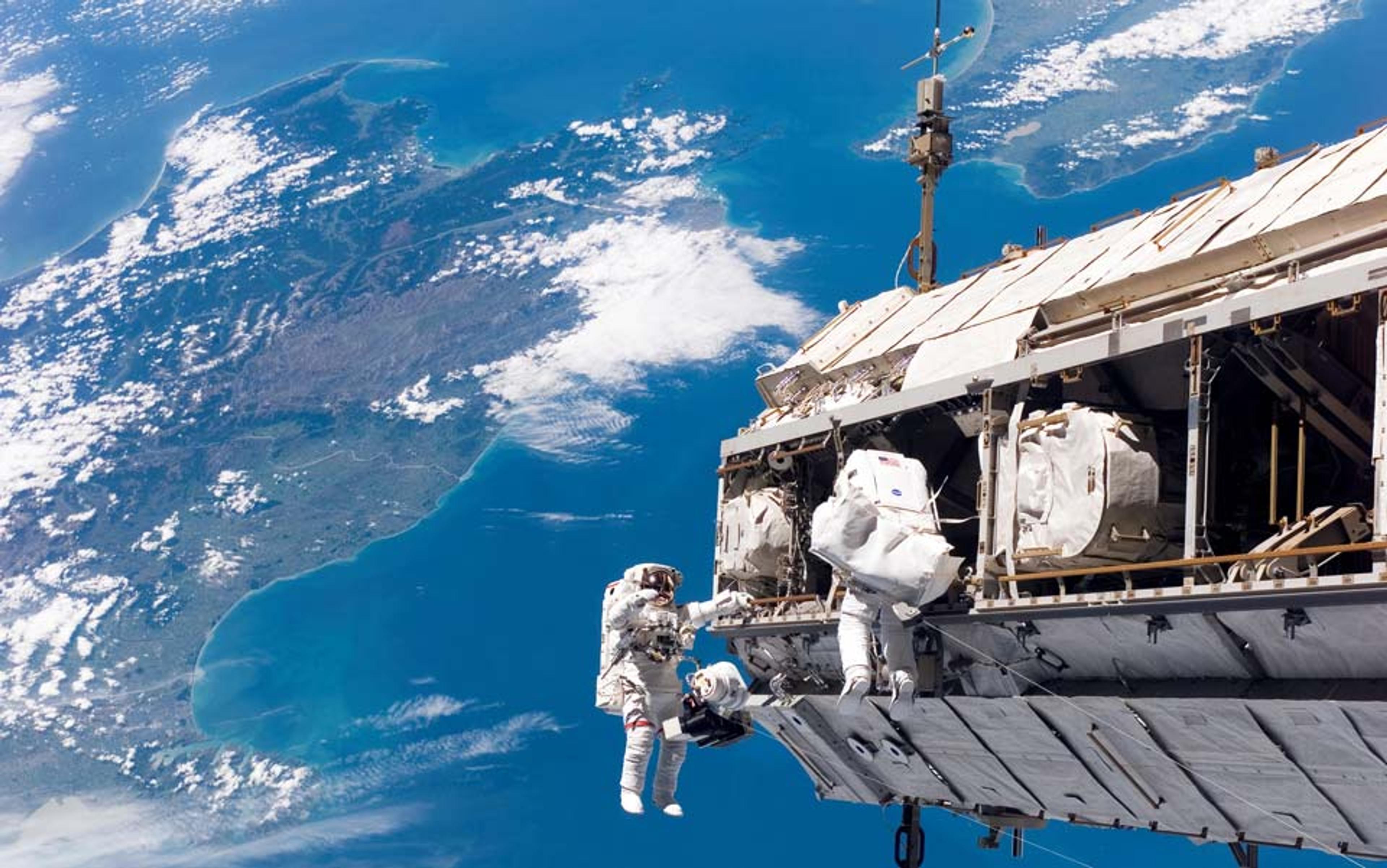 Two astronauts working on the International Space Station with Earth and its blue oceans in the background.