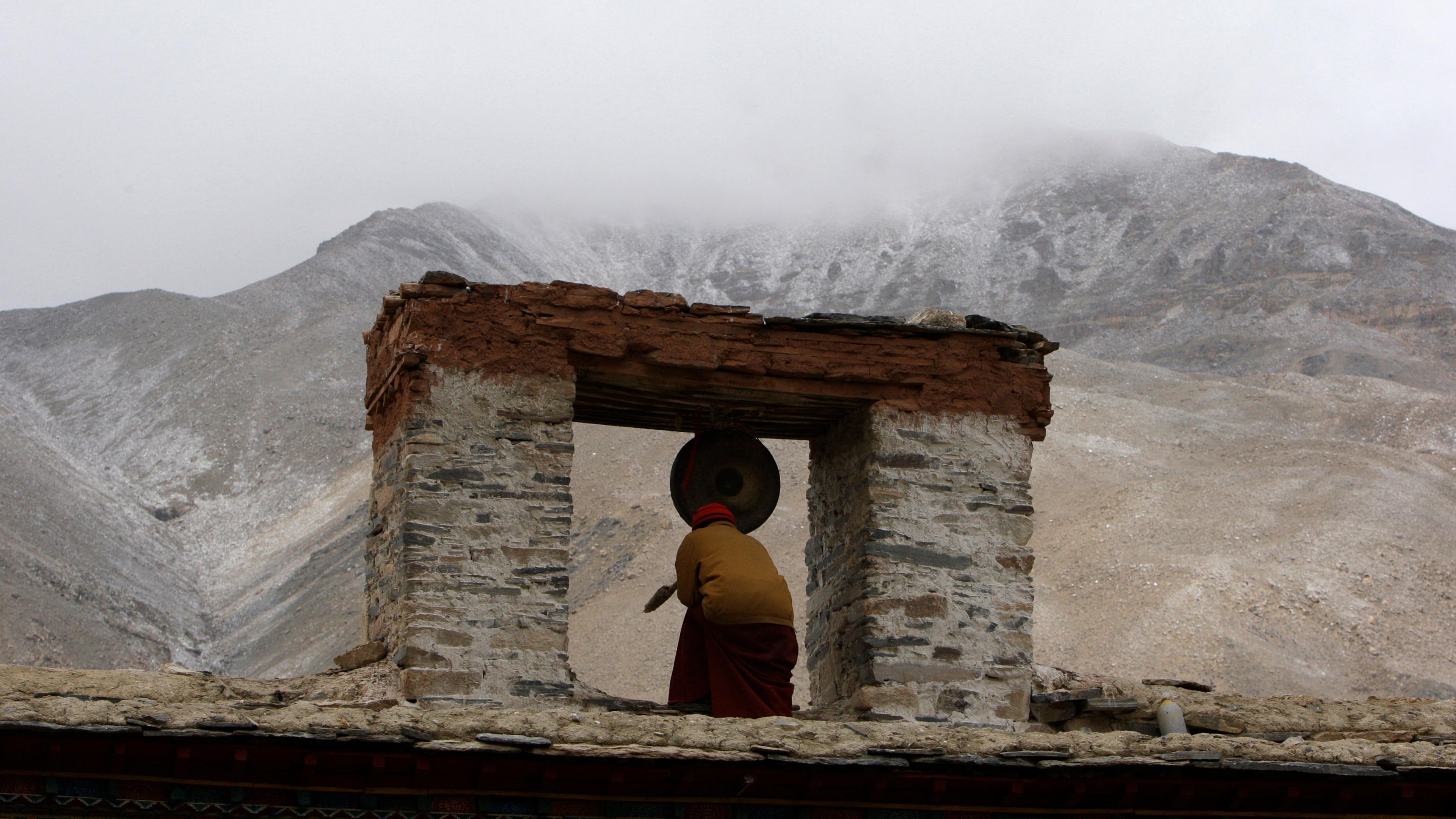Photo of a monk striking a large gong in an open stone structure with misty mountains in the background.