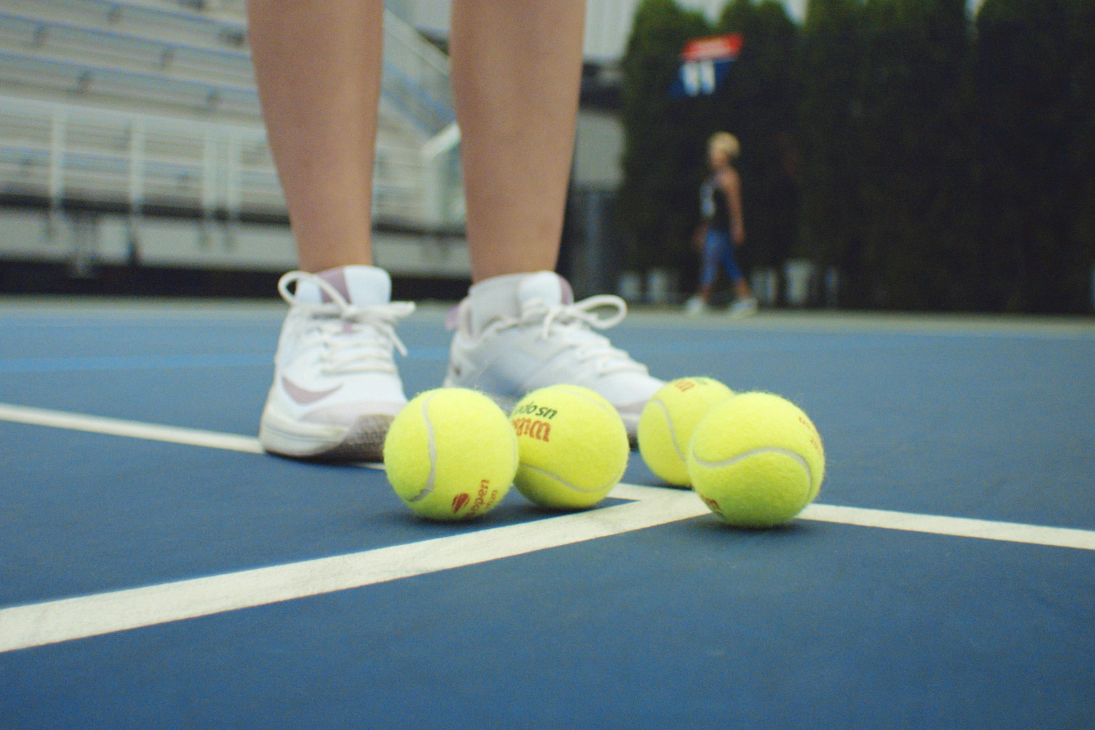 Close-up of feet in white trainers near four tennis balls on a blue tennis court, with blurred background of a few people.