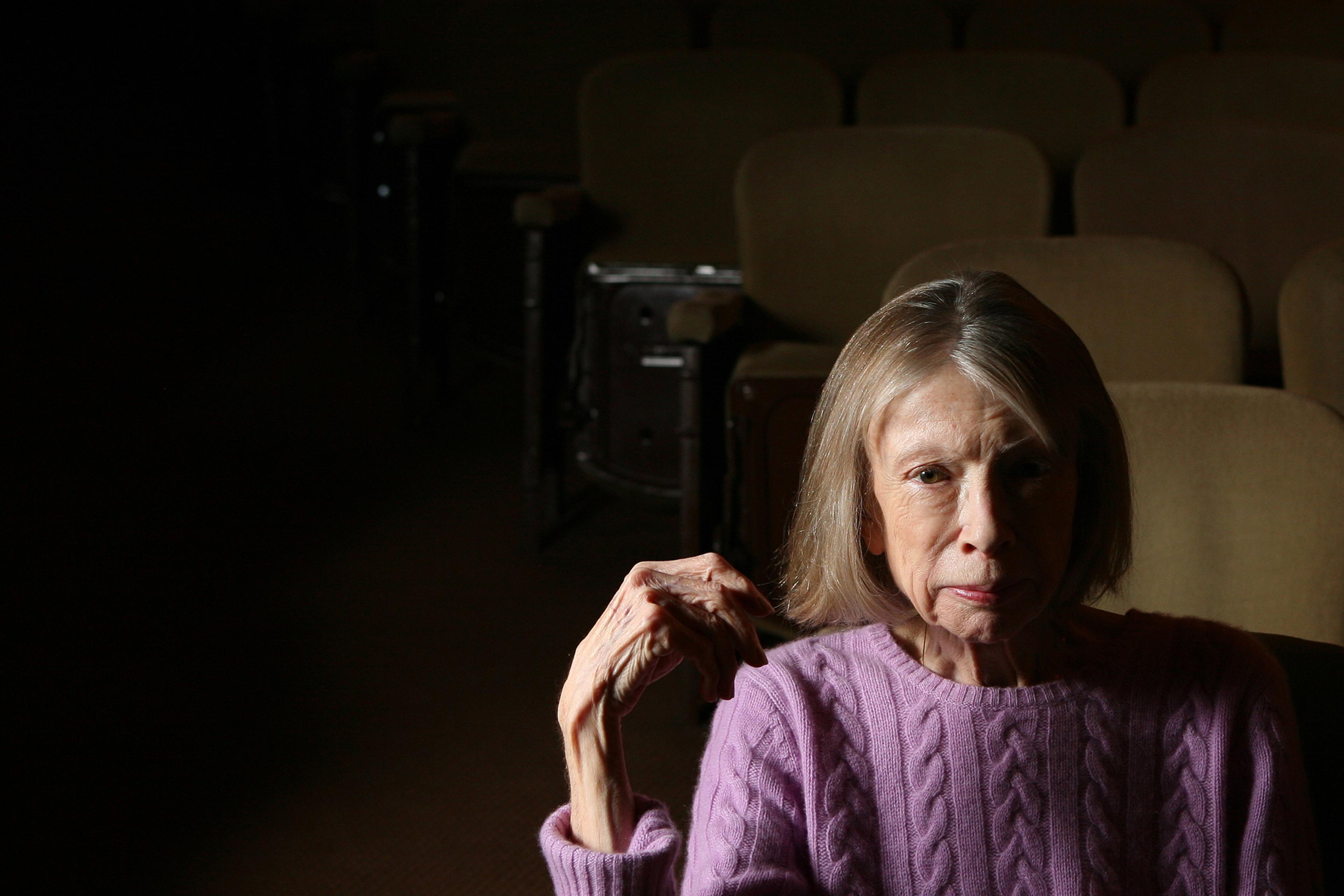 Photo of an older woman in a purple jumper sitting alone in dimly lit theatre seats.