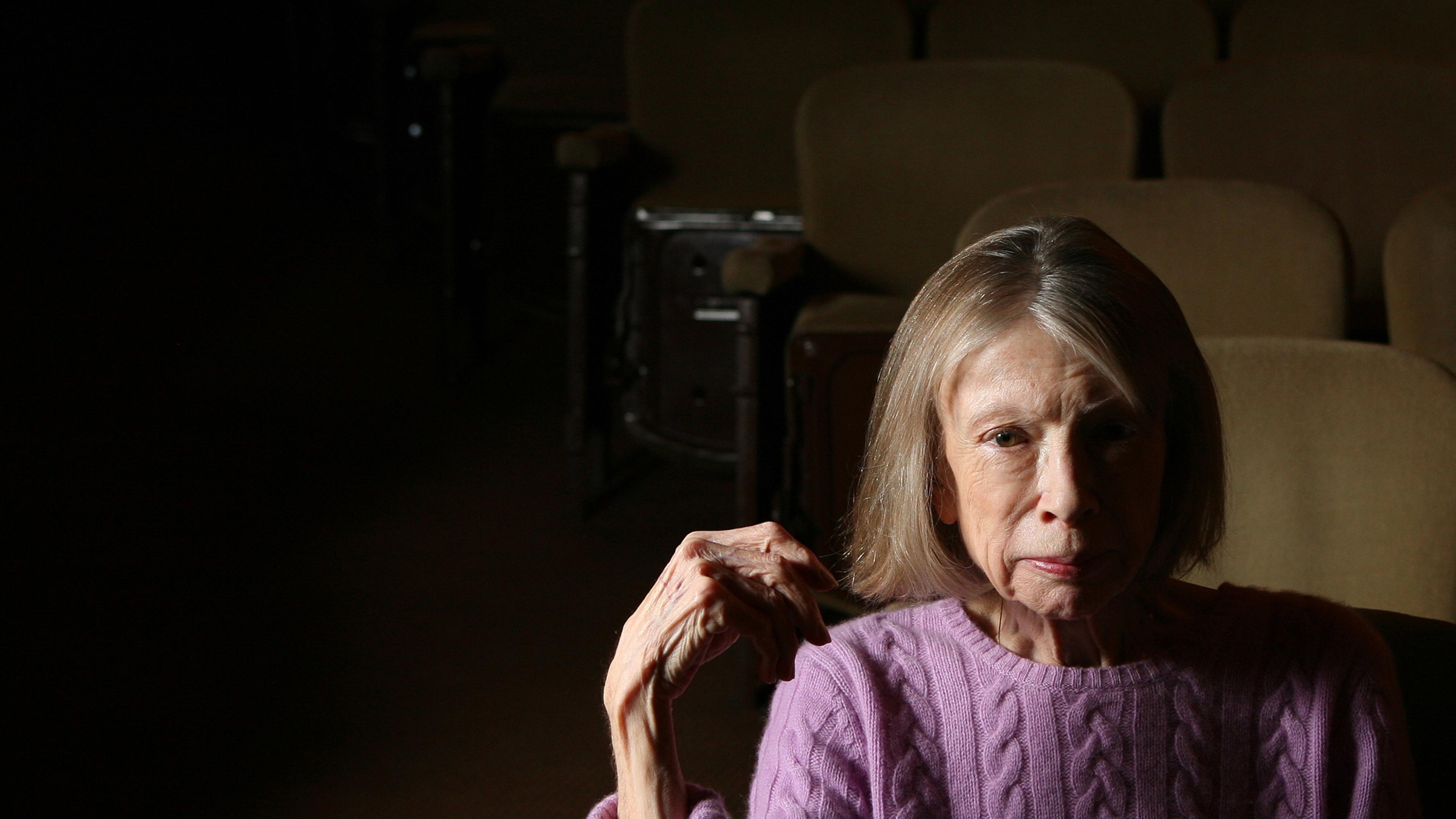 Photo of an older woman in a purple jumper sitting alone in dimly lit theatre seats.