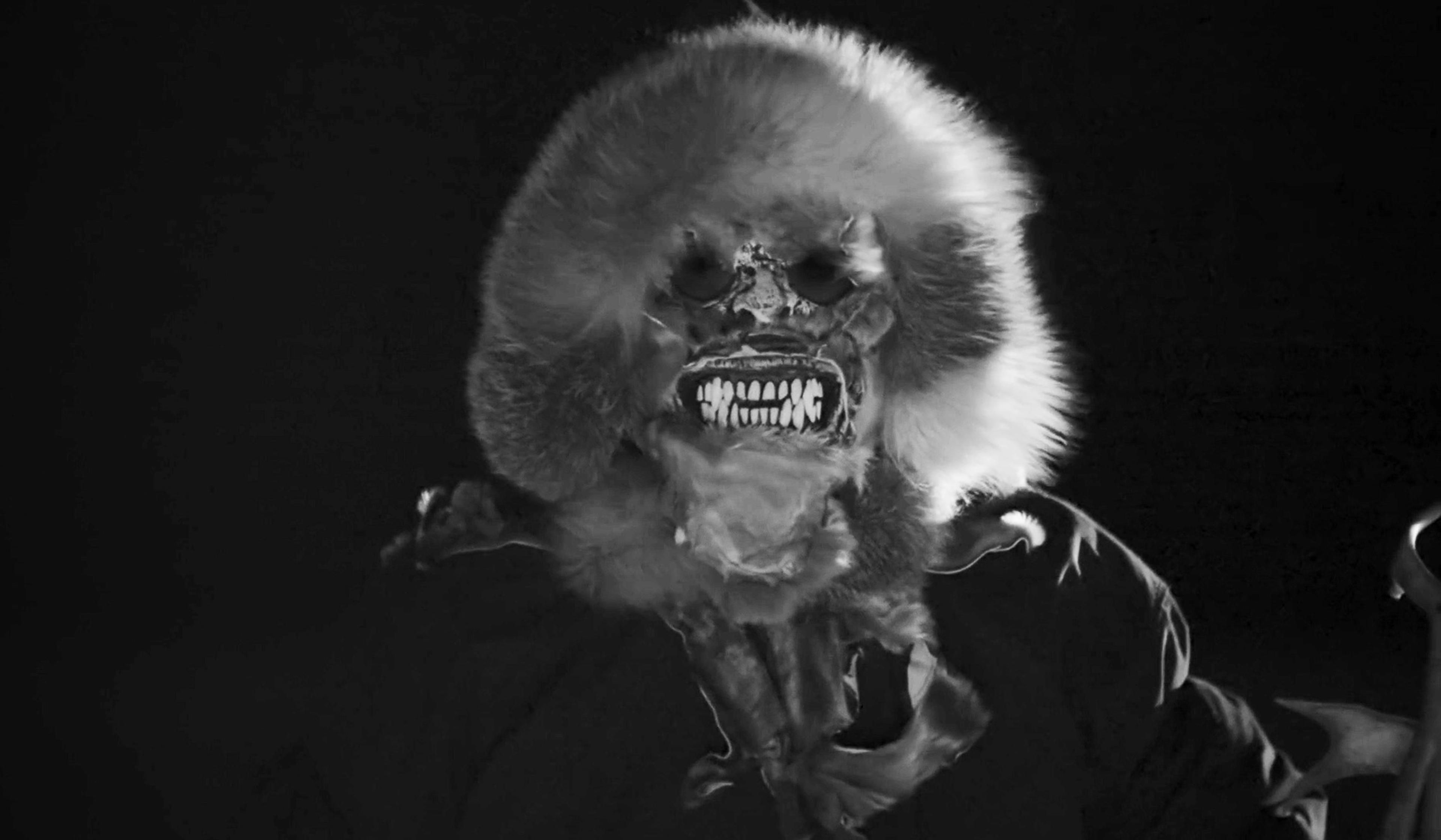 A scary figure in a fur-lined hood with a skeletal face and sharp teeth, in black and white against a dark background.