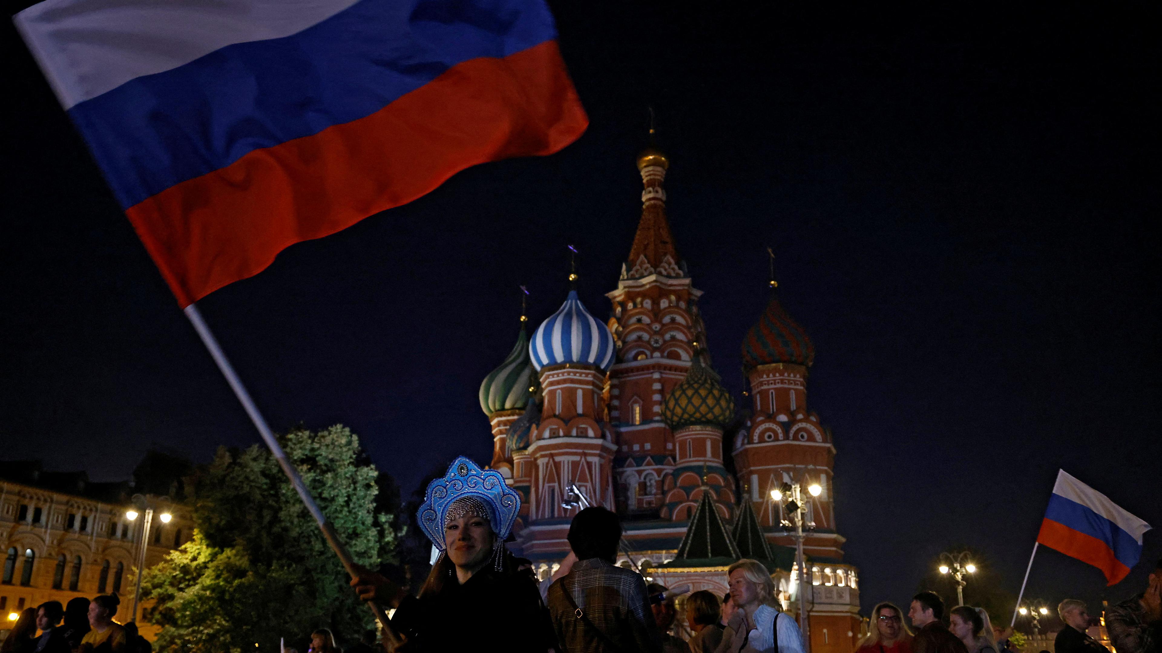 People at night in front of St Basil’s Cathedral in Moscow with a woman waving a large Russian flag in the foreground.