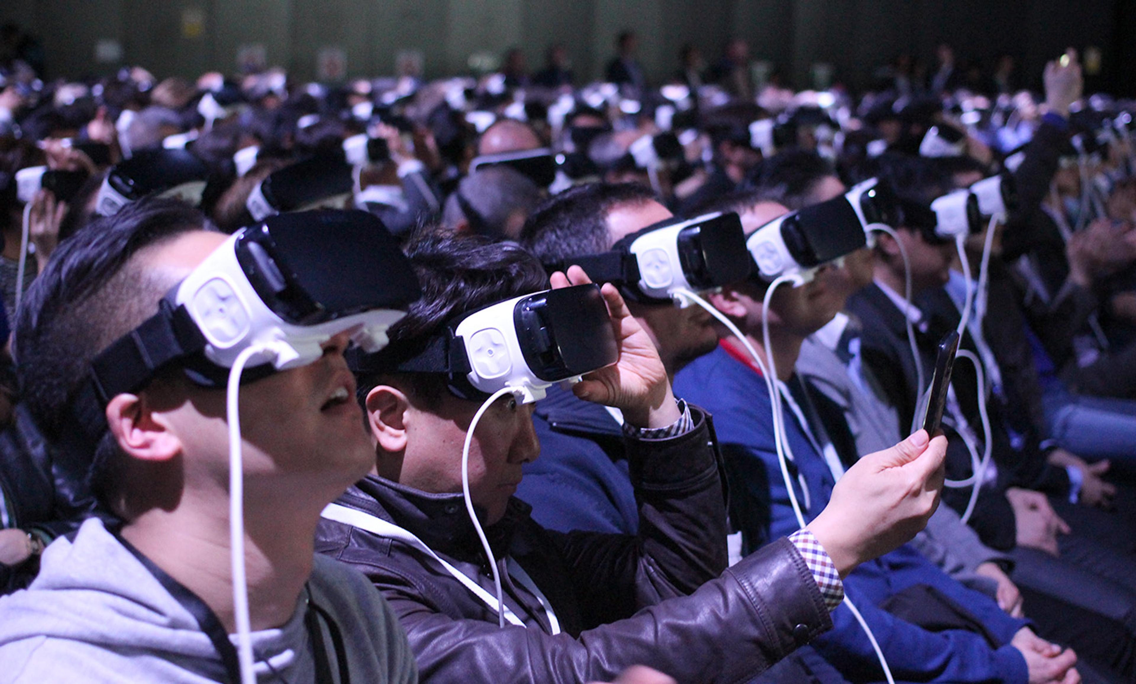 A large group of people seated wearing virtual reality headsets indoors, some holding controllers or smartphones.