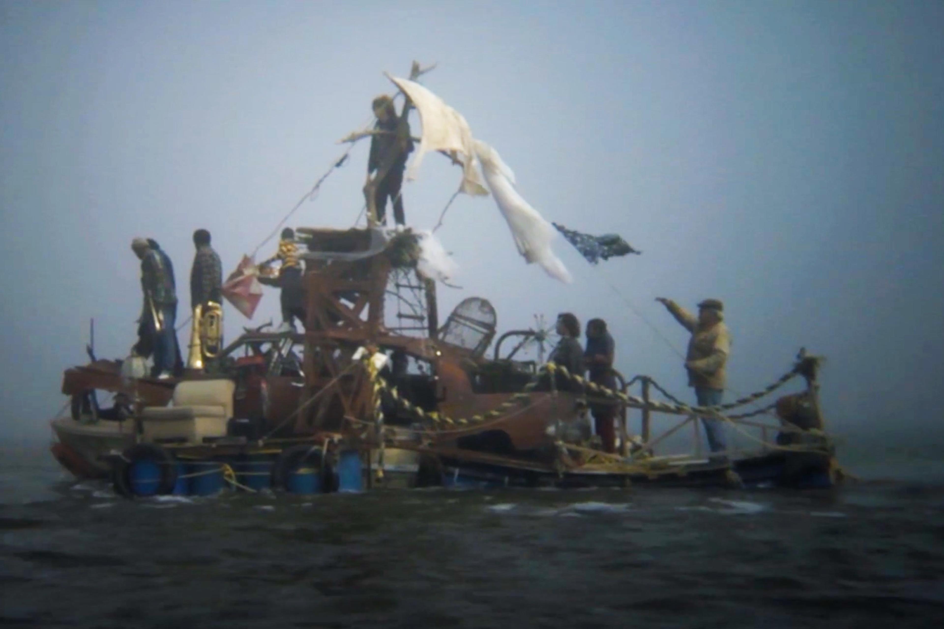 People on a makeshift raft, constructed from various items and barrels, floating on water under a grey, foggy sky.