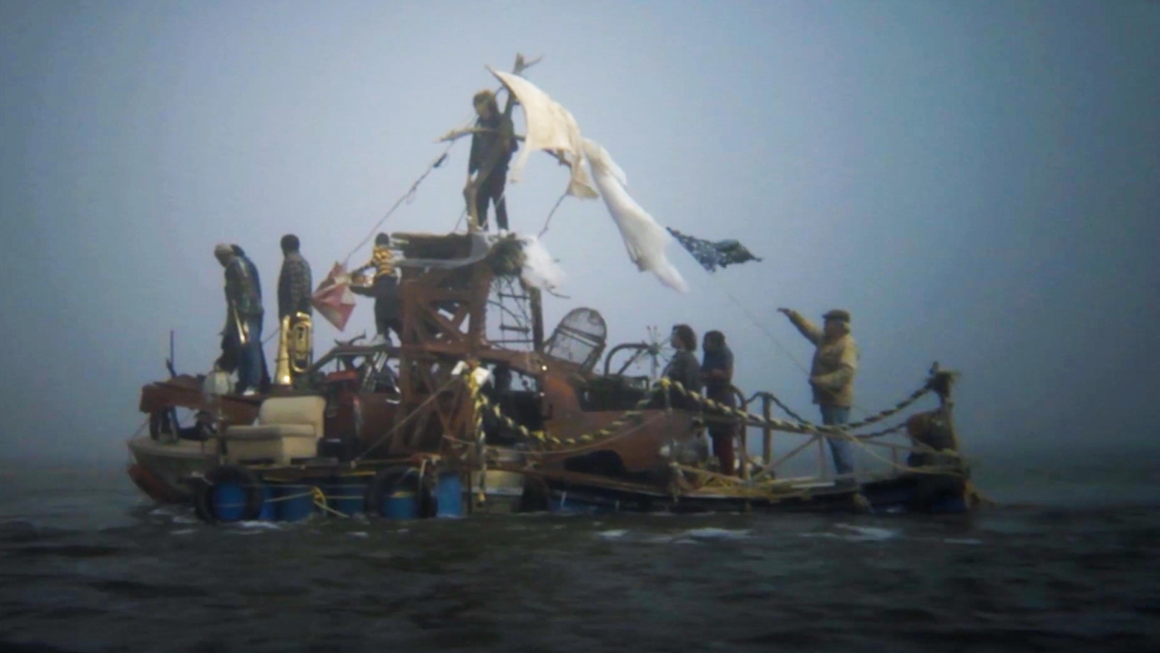People on a makeshift raft, constructed from various items and barrels, floating on water under a grey, foggy sky.
