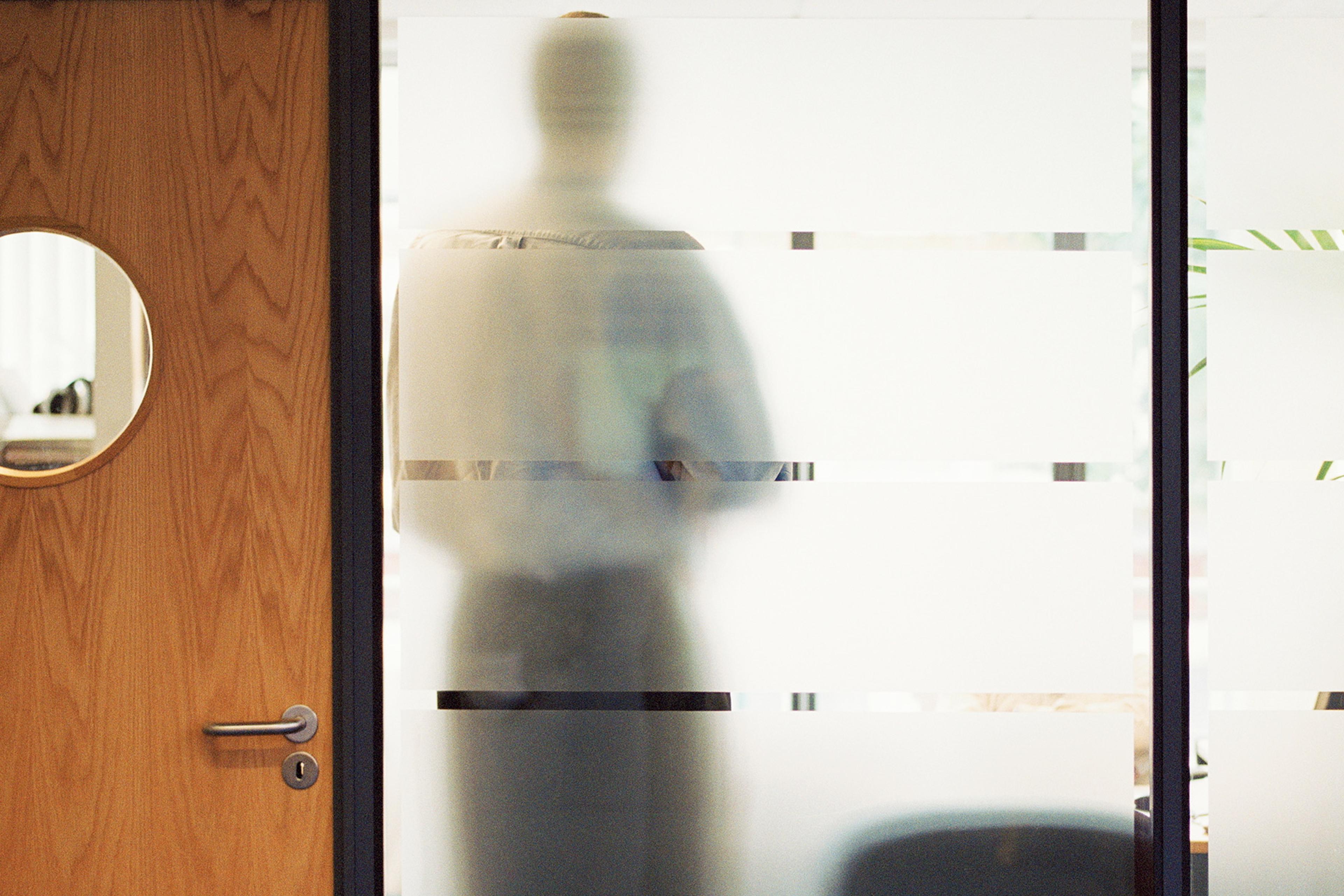 Photo of a person standing behind frosted glass in an office, obscured by the glass’s patterns.